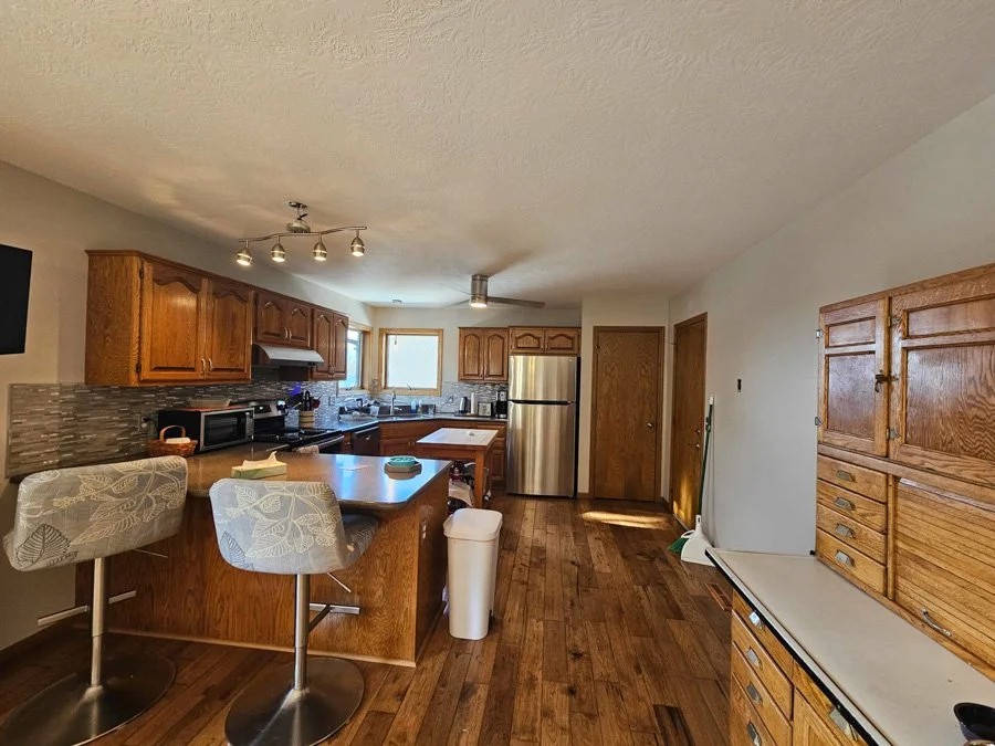 Kitchen with wooden cabinets, stainless steel refrigerator, and island with two patterned chairs in the foreground. Wood flooring and a small window above the sink. Wooden armoire on the right side.