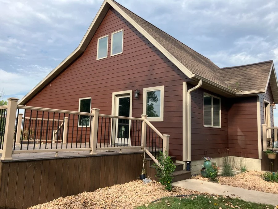 A house with brown siding, a deck with railing, and a gabled roof under a cloudy sky.