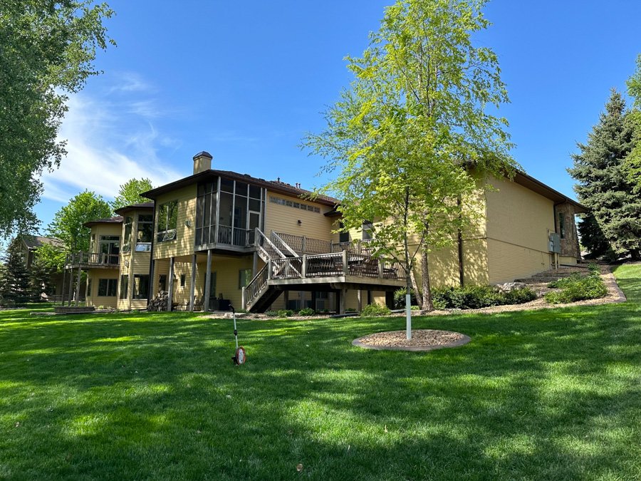 A large, two-story yellow house with a deck and screened porch, surrounded by green grass, trees, and shrubs under a bright blue sky.