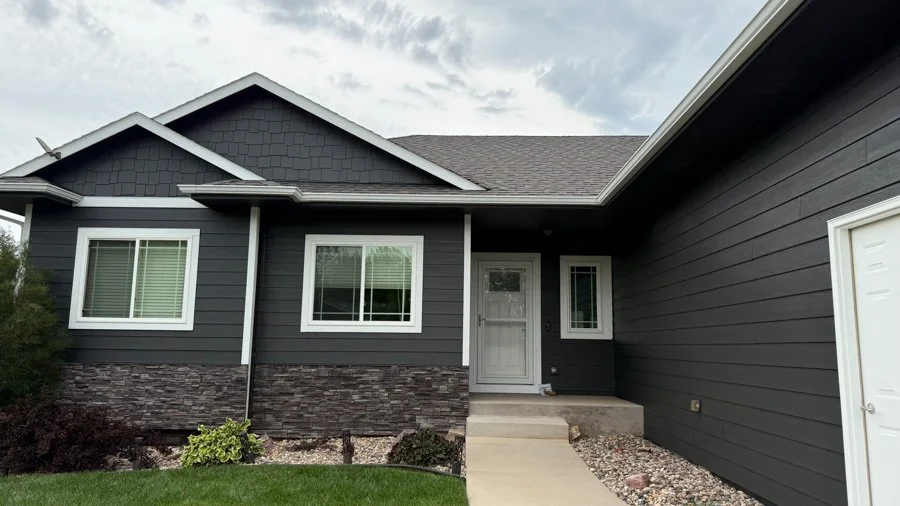 Front view of a modern house with dark gray siding, white trim, and a stone facade at the base, including a white front door and a small porch with steps, and a partly cloudy sky overhead.