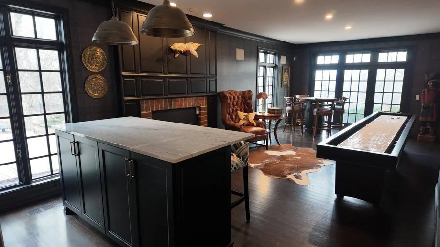 A living room with dark walls, hardwood floors, large windows, and modern furniture. It features a marble-top kitchen island, a brick fireplace, a high-back armchair, a cowhide rug, and a bar table with chairs. There are decorative plates on the wall