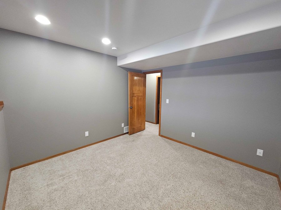 Empty room with gray walls, beige carpet, wooden trim, door open to another room, ceiling lights, and electrical outlets.