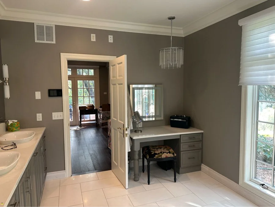 A bathroom with gray walls, a double sink vanity, a doorway leading to an adjacent room with a table and chairs, and a window with white blinds letting in natural light.