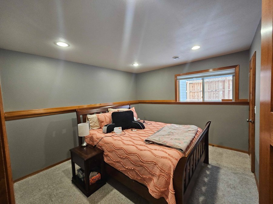 A bedroom with a wooden bed, pink bedspread, and several pillows. There is a nightstand with a lamp and some items, and a window showing a wooden fence outside.