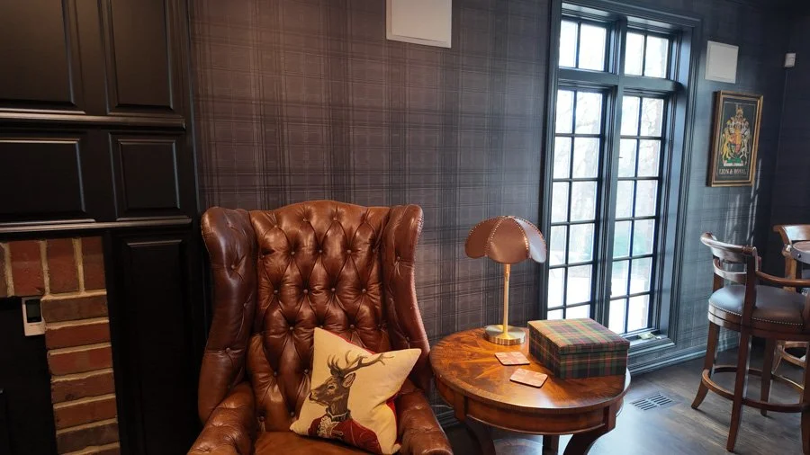 Cozy interior with a brown leather wingback chair, a plaid throw pillow, a small wooden side table with a decorative lamp, a window with grid panes, and a wall with framed artwork.