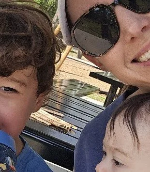 A woman and two children outdoors, smiling, with a bench and trees in the background.