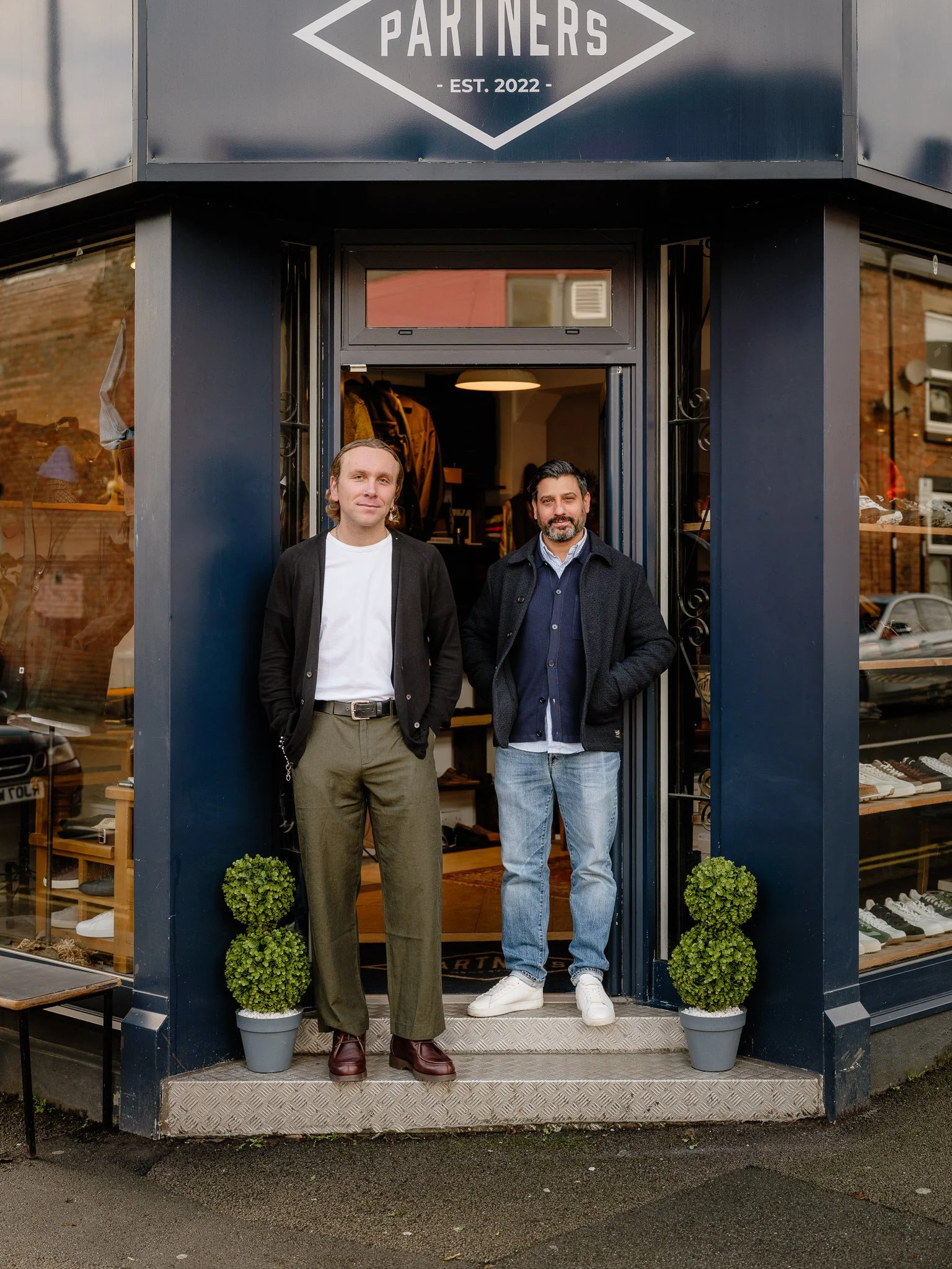 Two men standing in front of a clothing store, named 'Partners,' established in 2022, with plants on either side of the entrance.