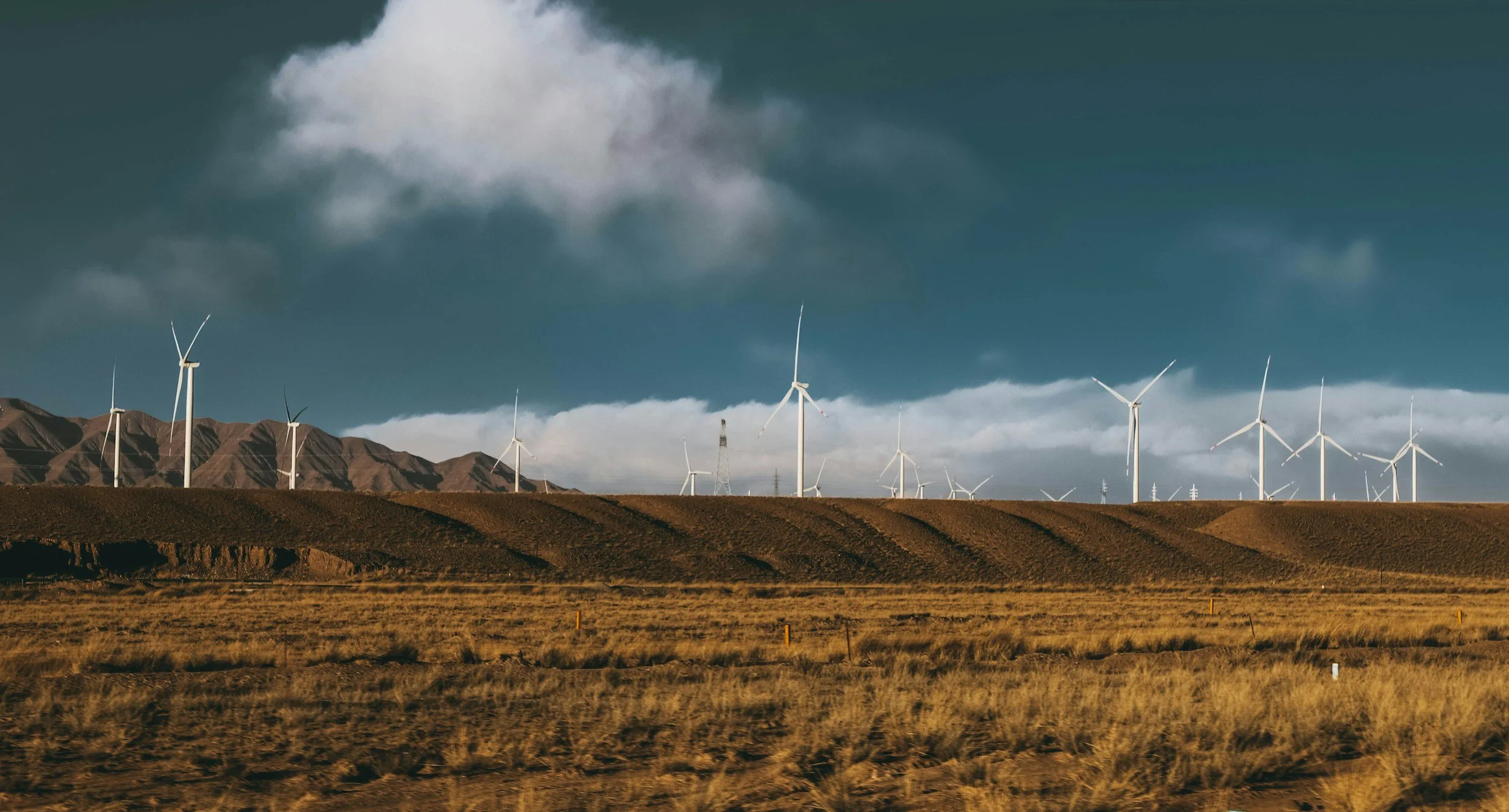 A landscape of a wind farm with multiple wind turbines on rolling hills, with mountains in the background, dark clouds in the sky, and dry grass in the foreground.