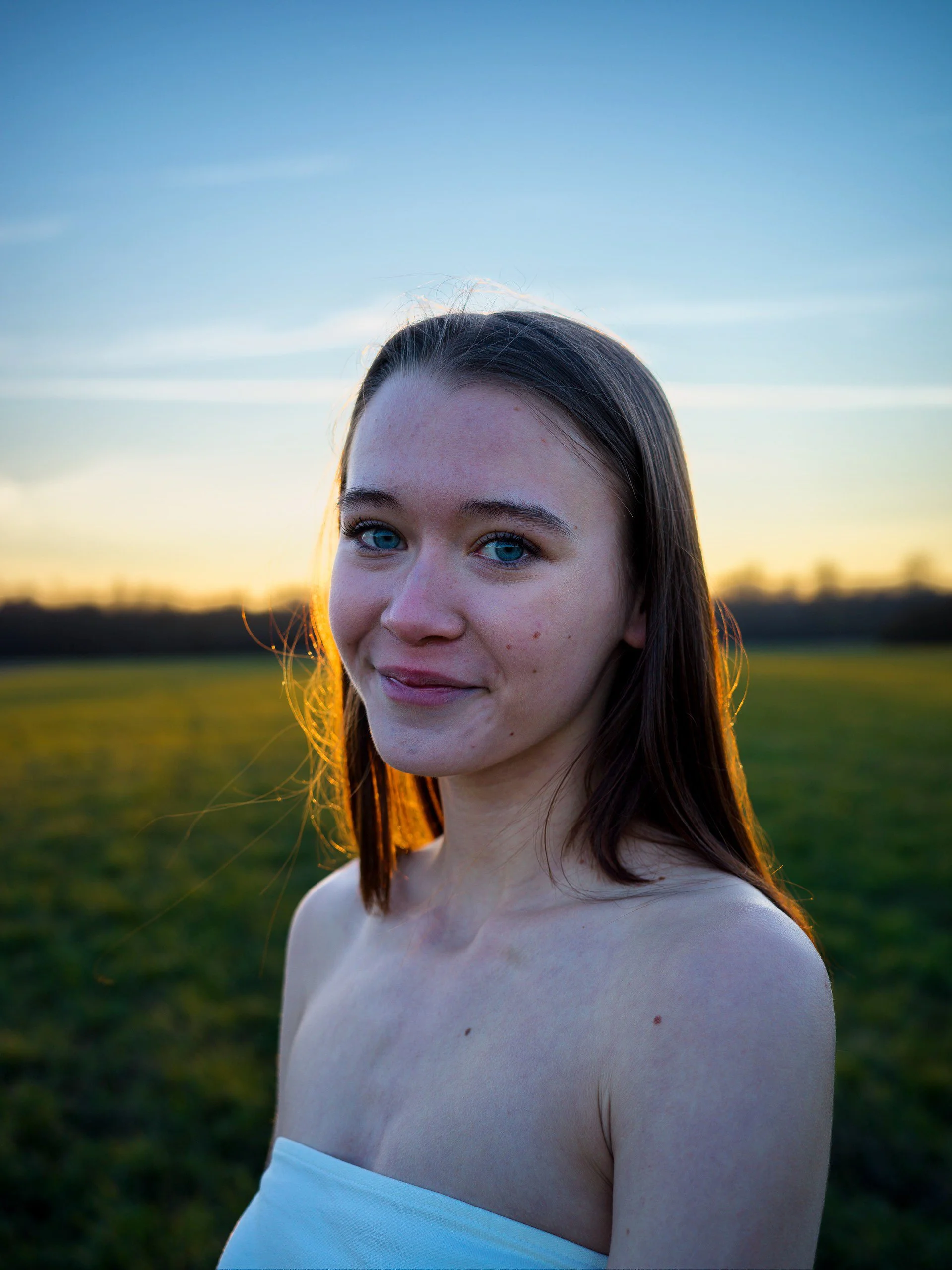 Junge Frau mit braunen Haaren und blauen Augen im Freien bei Sonnenuntergang in einer offenen Landschaft.