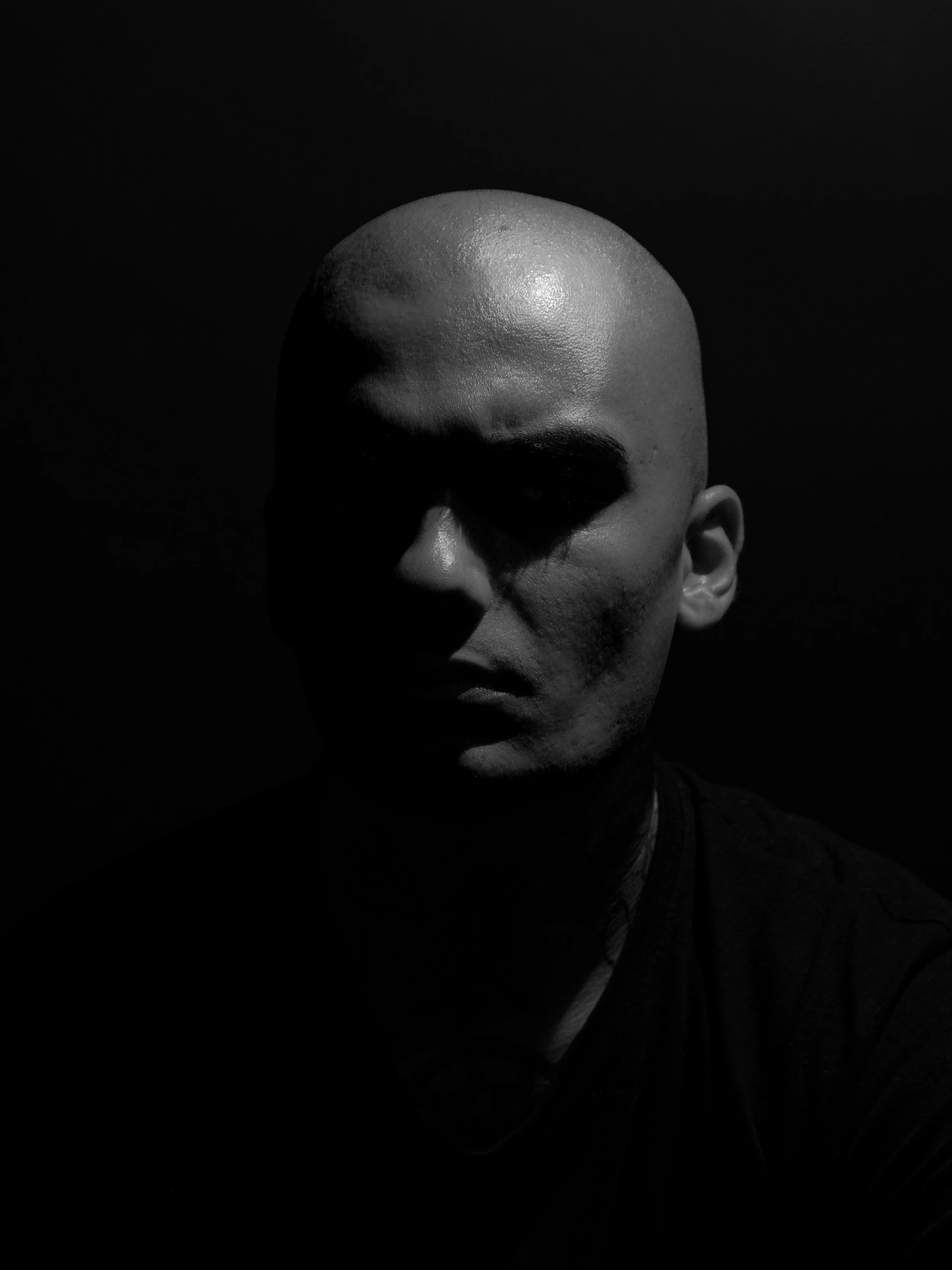 Schwarz-weiß-Fotografie einer Person mit Glatze, halboffenes Gesicht, düsteres Licht, dunkler Hintergrund.