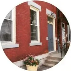 Red brick building with two large windows and a dark door, with a flower pot on the stairs outside.