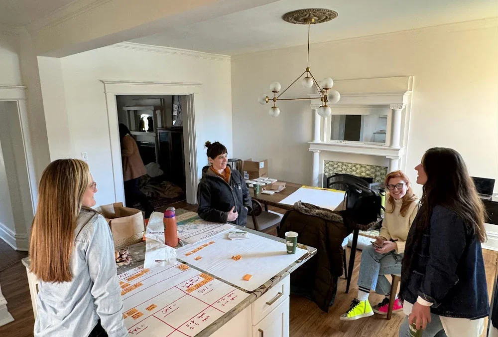 Four women in a kitchen or dining area engaged in conversation, with a table and paperwork in front of them.