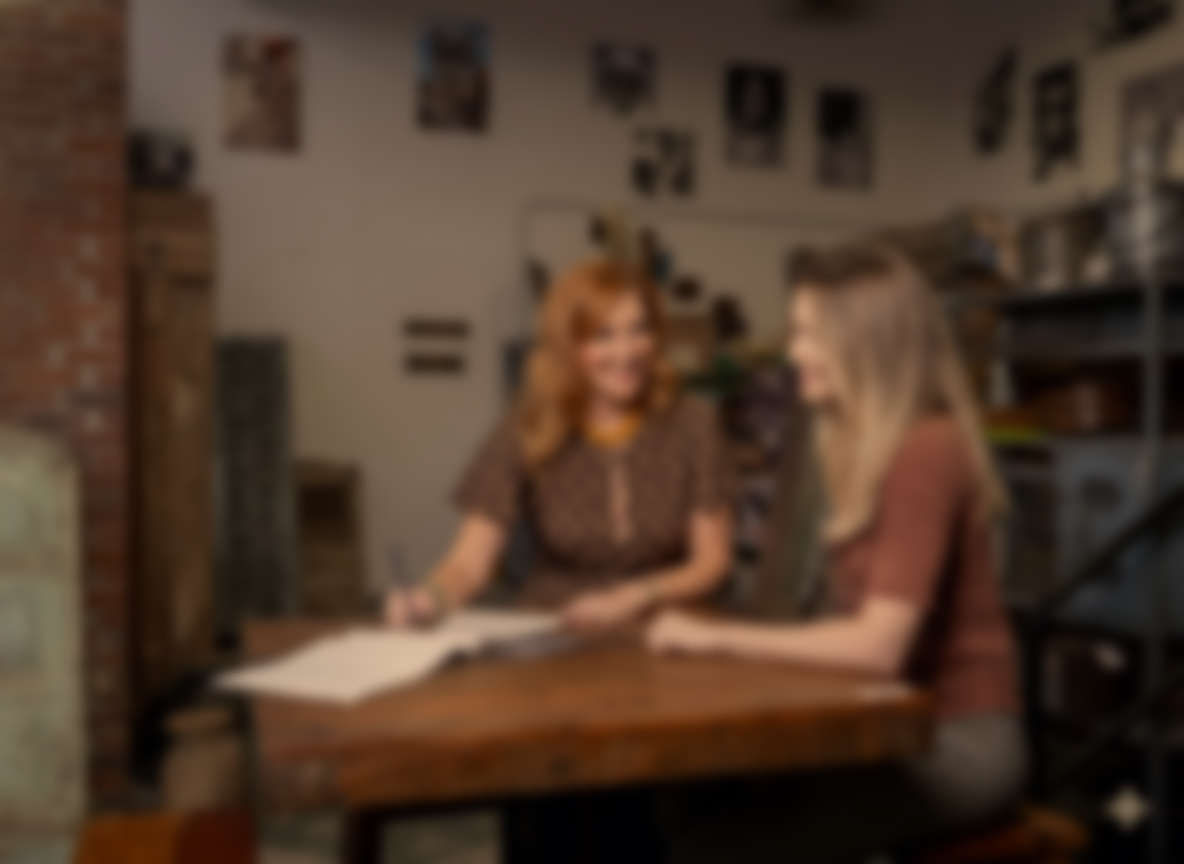 Two women sitting at a wooden table in a cozy indoor space, smiling and having a conversation, with photographs on the wall behind them.
