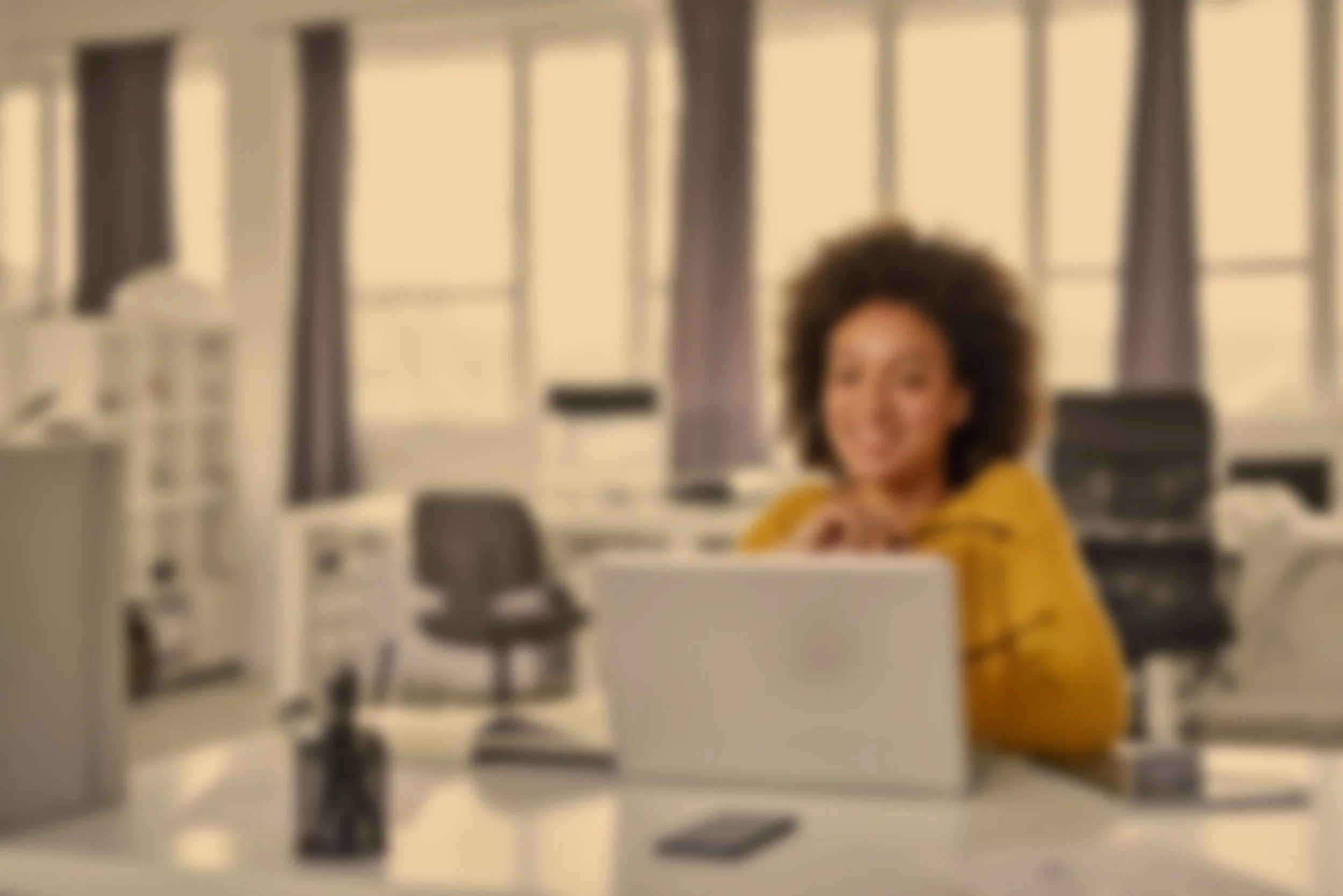 A woman with curly hair smiling at a desk in an office environment with large windows and curtains in the background.