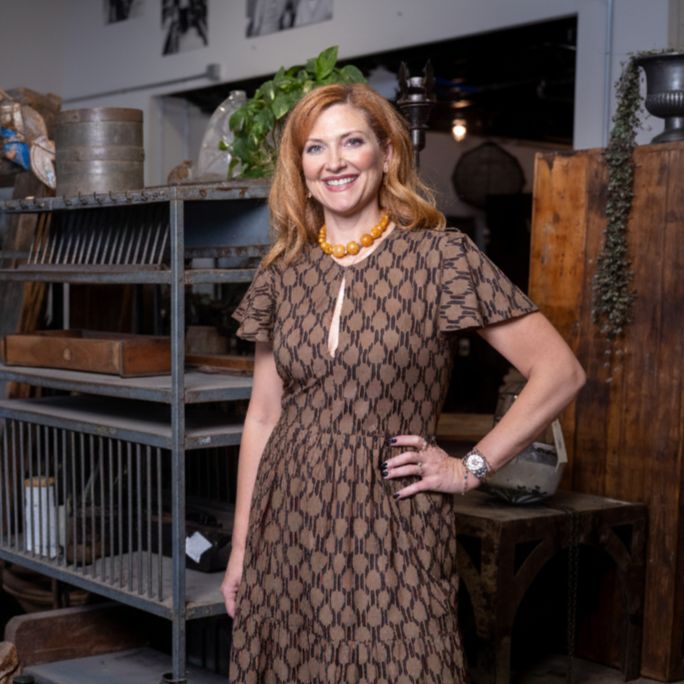 A woman with wavy, shoulder-length red hair, wearing a brown patterned dress with a keyhole neckline, a yellow beaded necklace, a watch, and a ring, standing indoors with a rustic, eclectic decor background.