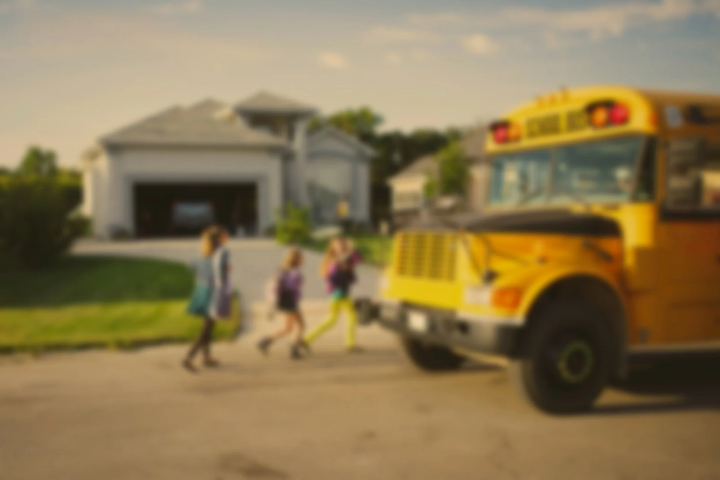 Blurry image of three students walking past a yellow school bus in a suburban neighborhood with houses and a garage in the background.