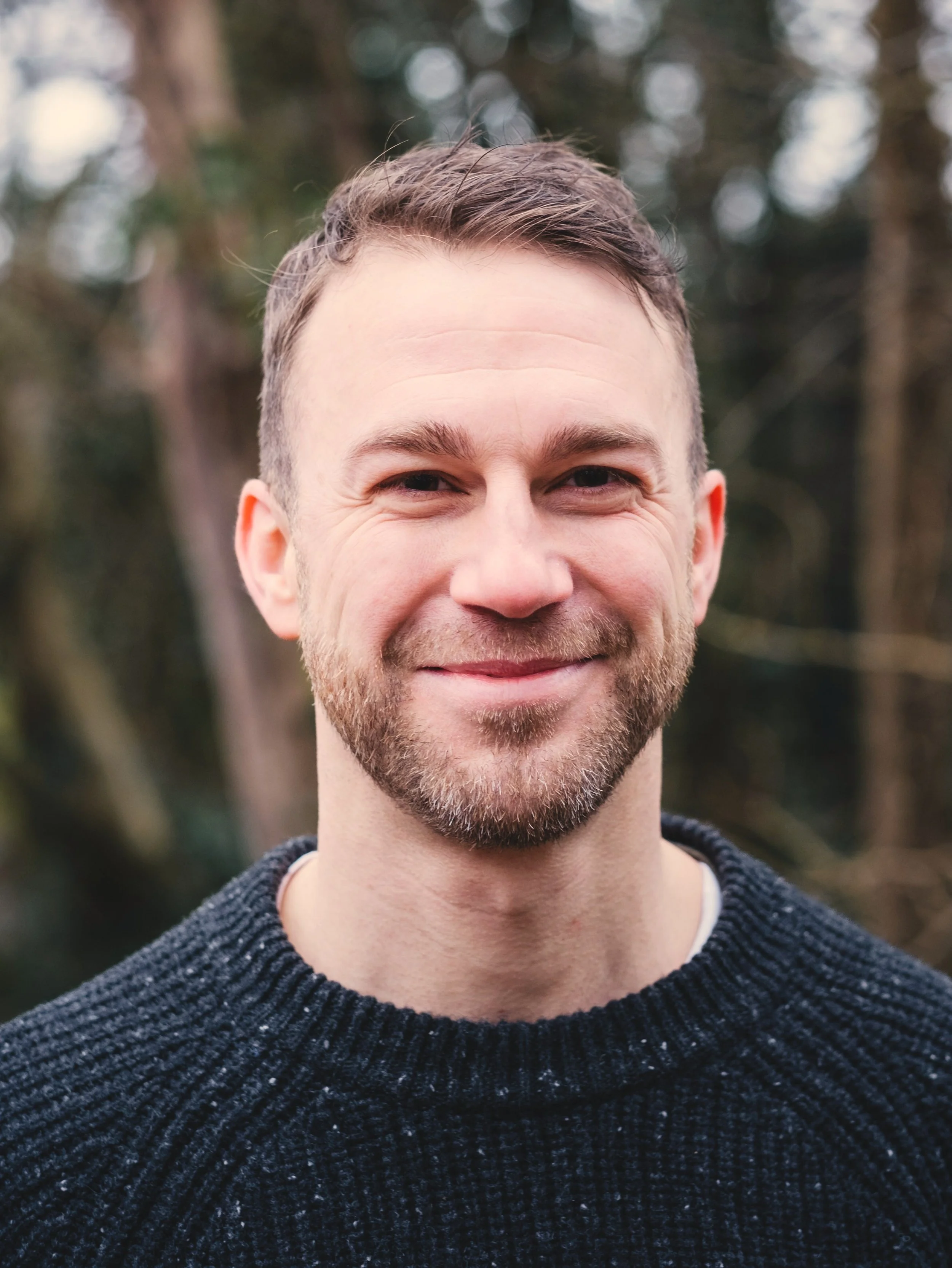 Close-up portrait of a smiling man with short brown hair and a beard, wearing a black sweater, outdoors with blurred trees in the background.