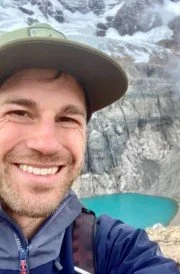 A smiling man wearing a hat takes a selfie outdoors with a waterfall and turquoise lake in the background.