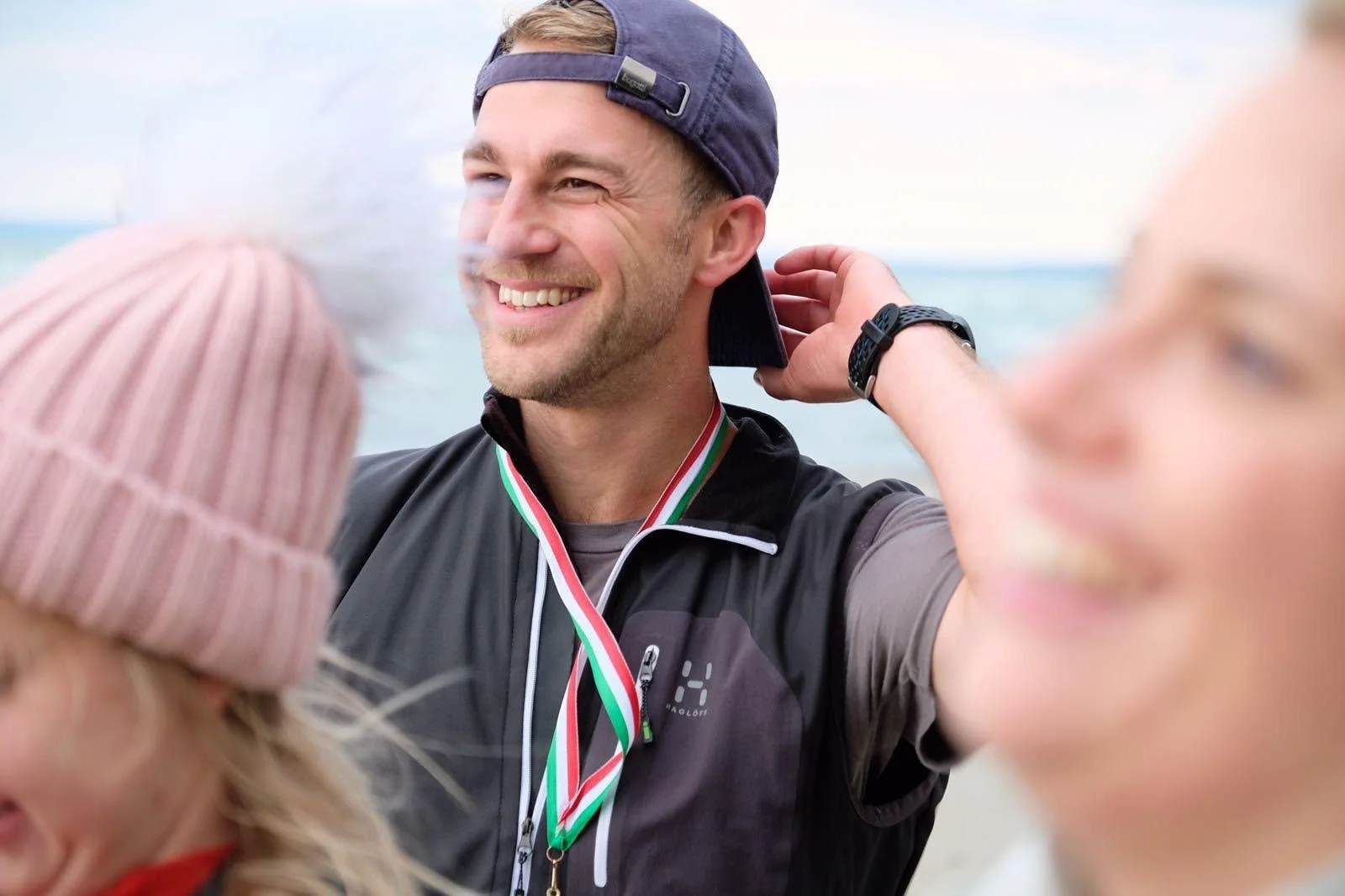 A man smiling as he wears a medal around his neck near the water, with two women in the foreground.