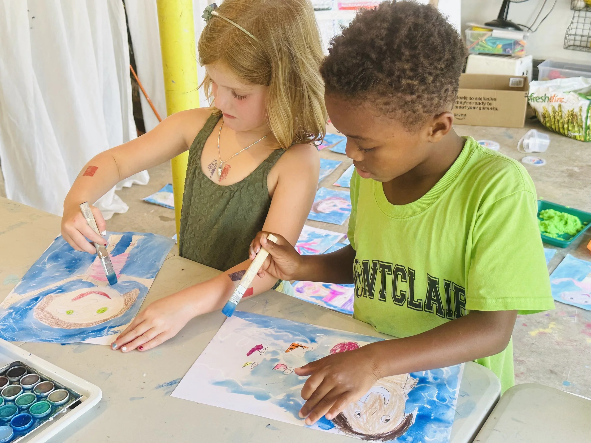Two children, a girl and a boy, painting at a table with watercolors. The girl is holding a paintbrush, and the boy is helping hold her artwork. They are creating colorful watercolor pictures, with various art supplies visible on the table and backgr