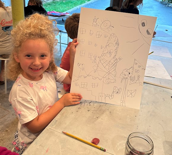 A young girl with curly blonde hair smiling and holding up a large drawing she made of various items, including a sun, a house, and several abstract shapes, in a classroom or art space.