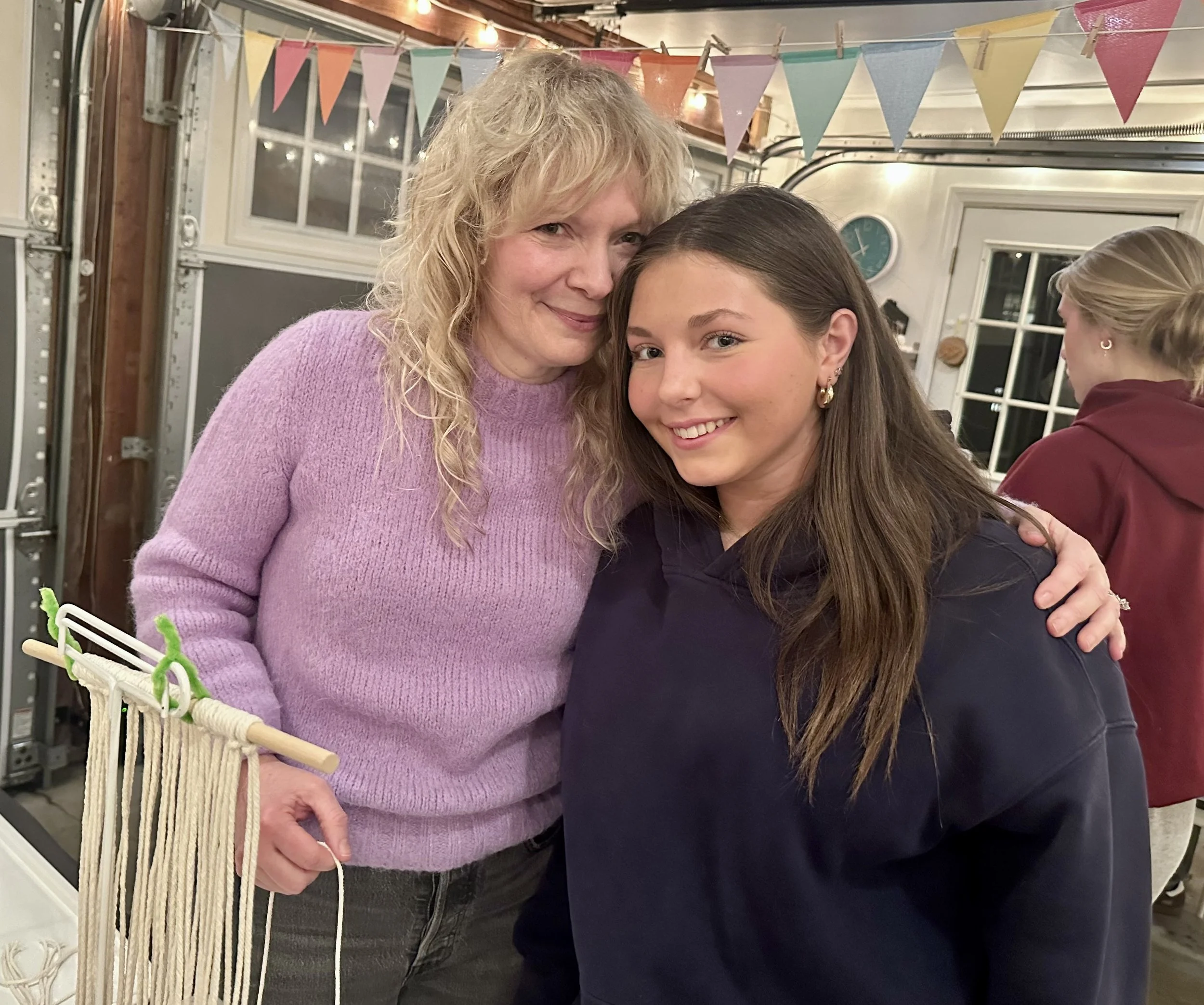 A woman with curly blonde hair wearing a pink sweater hugging a young woman with straight brown hair wearing a dark hoodie, both smiling, in a decorated indoor space.