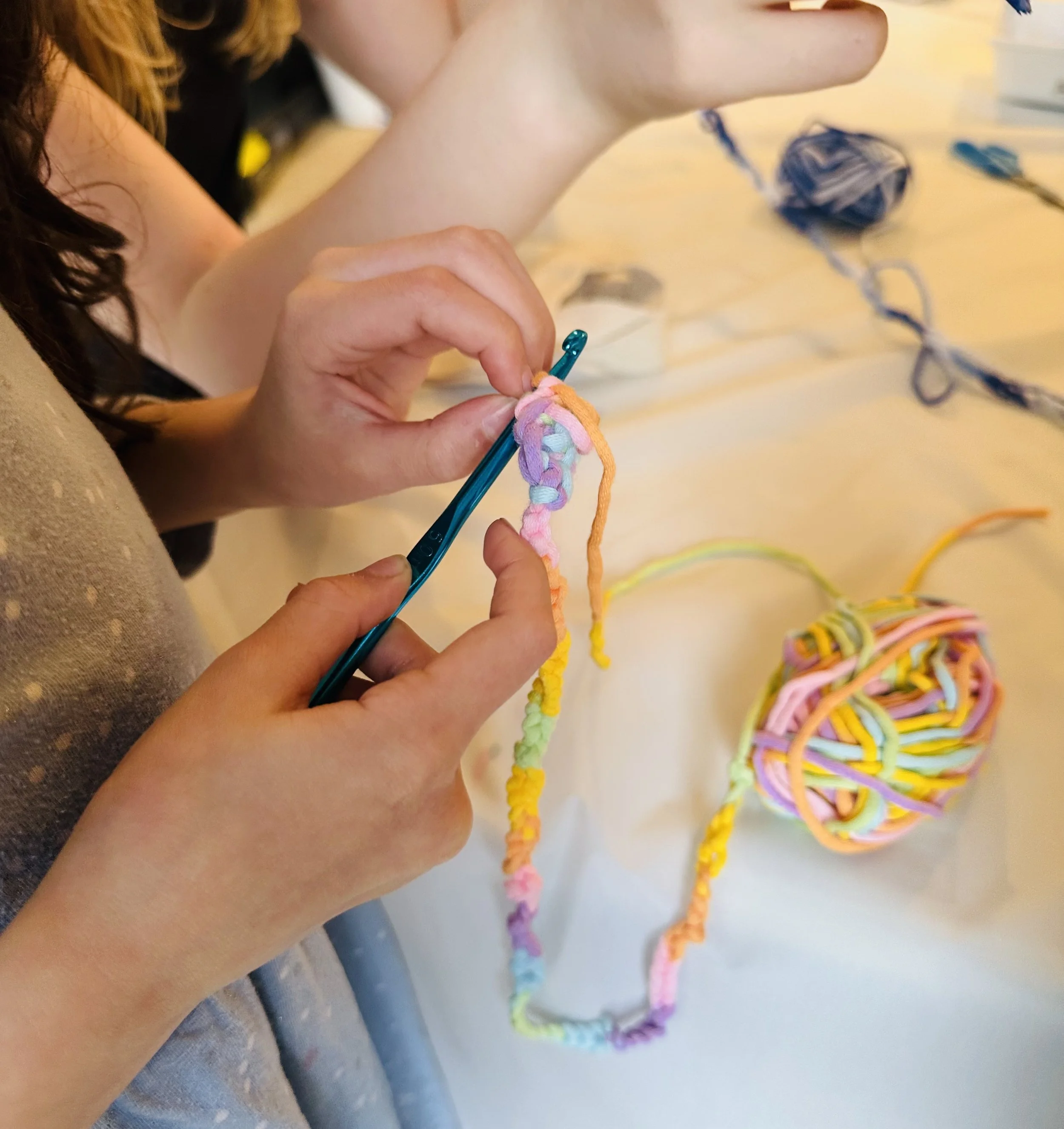 Person crocheting with colorful yarns at a craft table with a ball of multicolored yarn nearby.