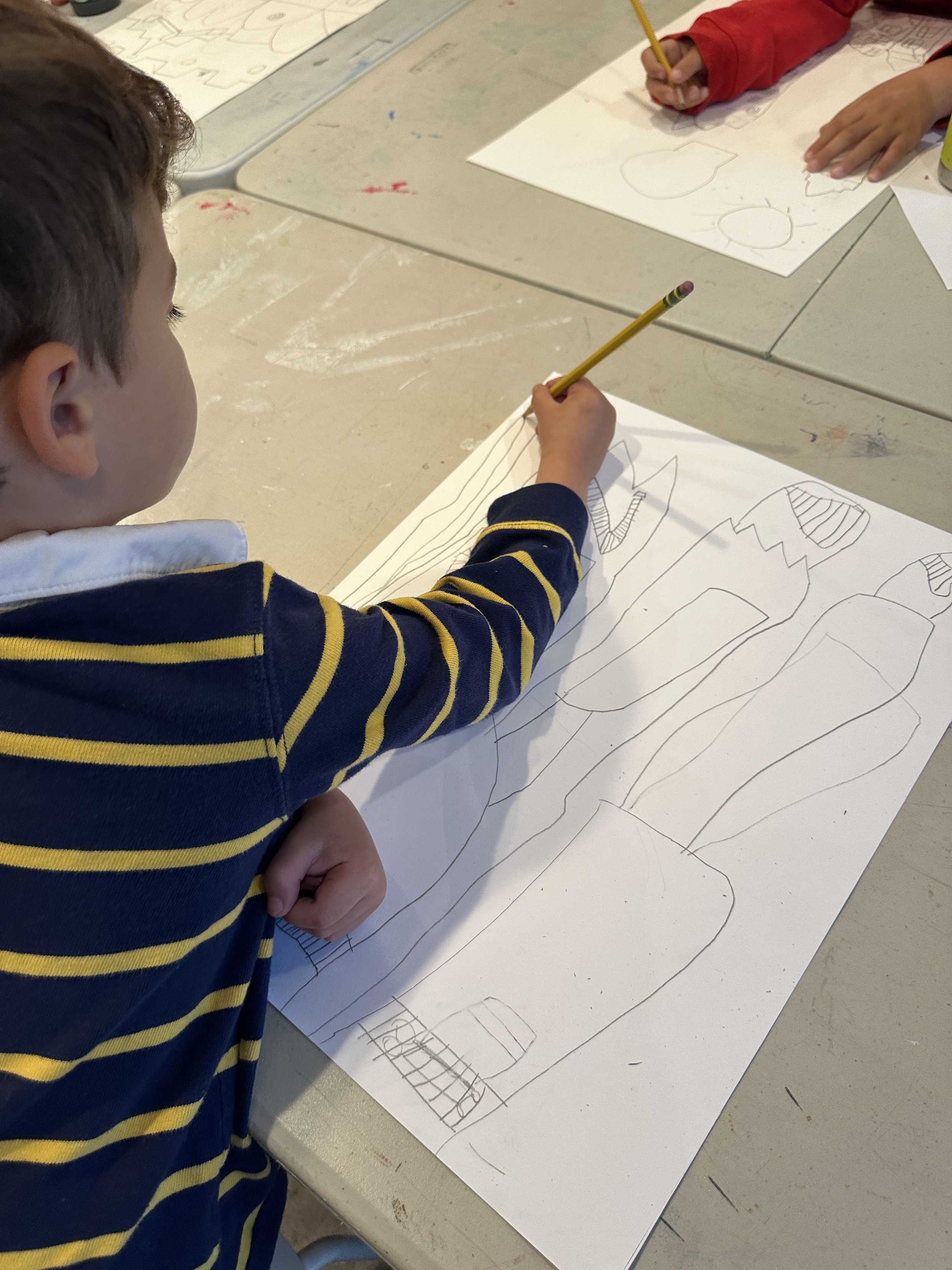 A young boy with short brown hair wearing a navy blue and yellow striped shirt drawing on a large sheet of paper with a pencil in a classroom setting.