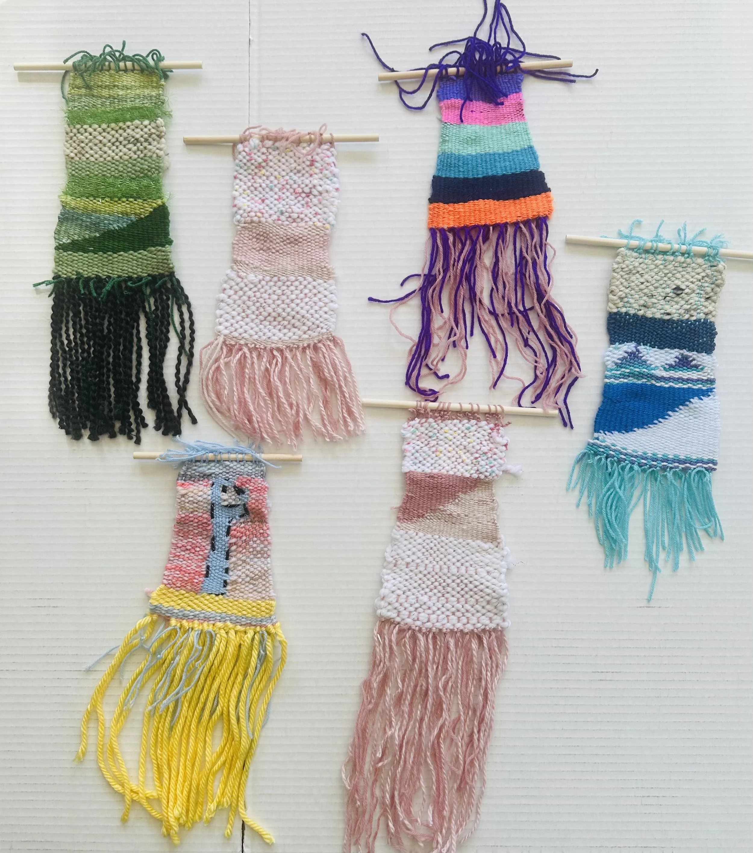 Seven small woven textile wall hangings with fringes, each featuring different colors and patterns, attached to horizontal rods or hanging by yarn.