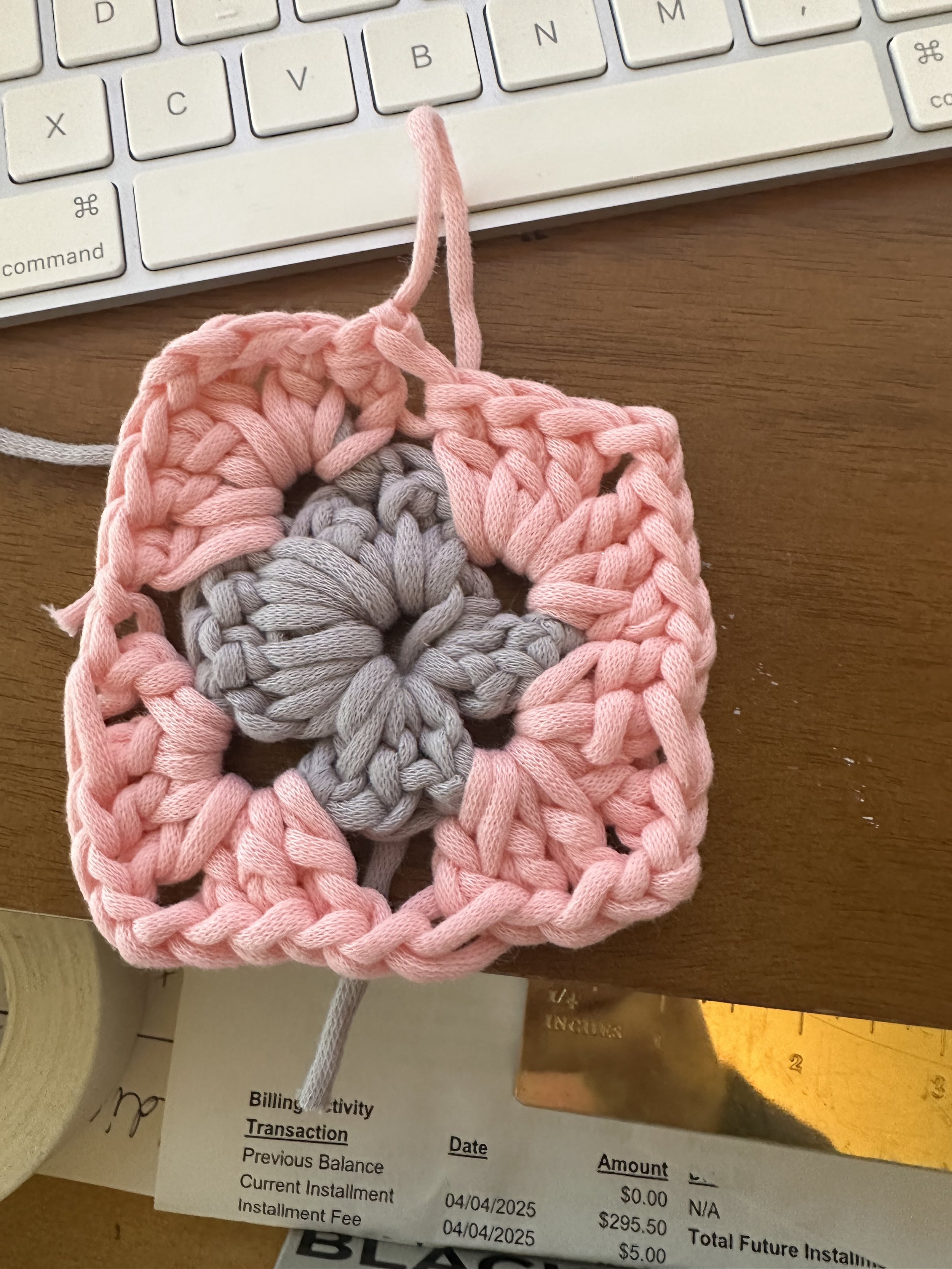A small handmade square gray crochet piece with a flower pattern in the center, surrounded by a pink crochet border, placed on a wooden desk near a keyboard and a bill receipt.