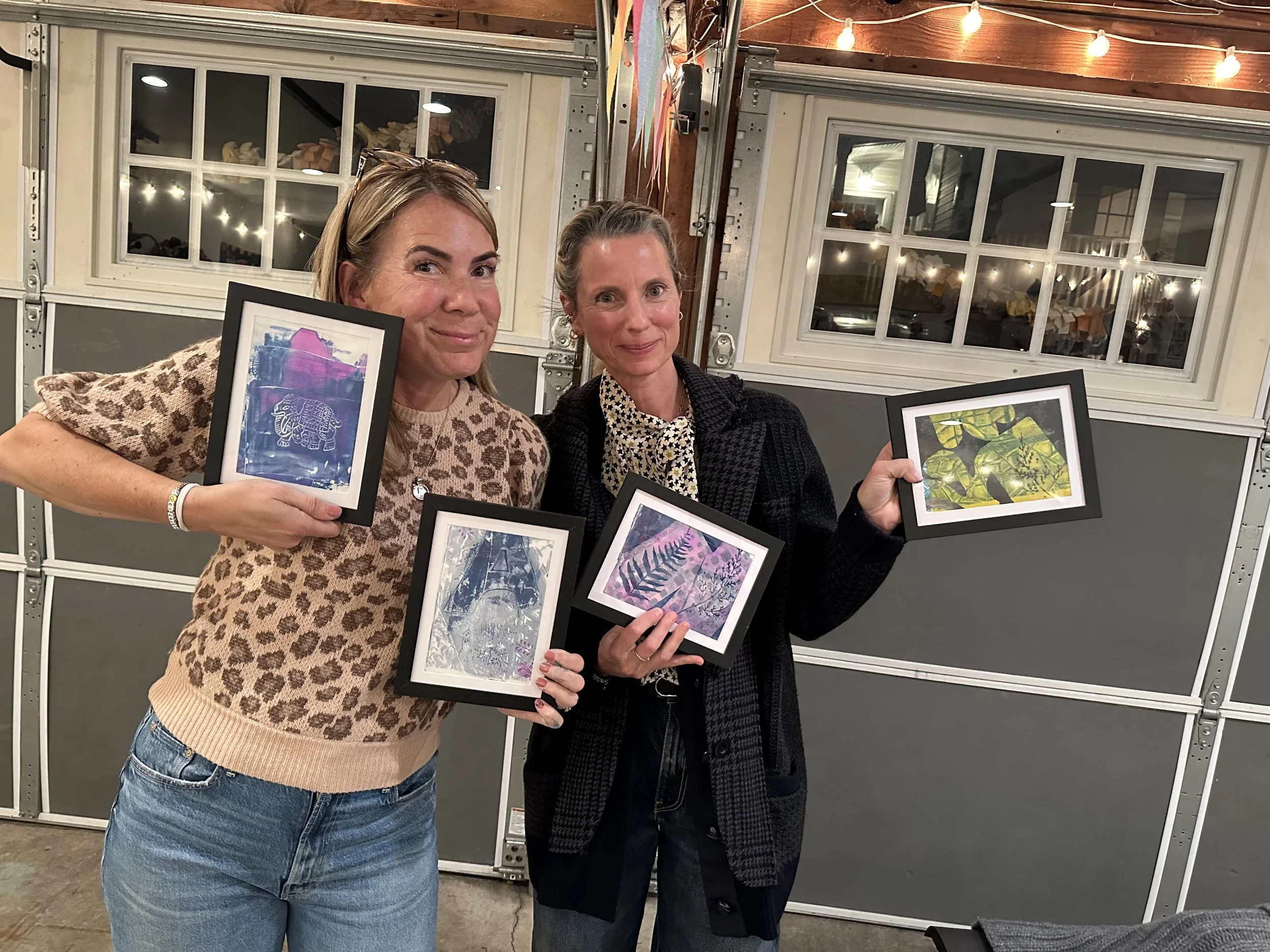 Two women standing inside a garage, each holding framed artwork featuring nature and wildlife images. The woman on the left has blonde hair, wearing a leopard print sweater and jeans, while the woman on the right has darker hair, wearing a dark coat 