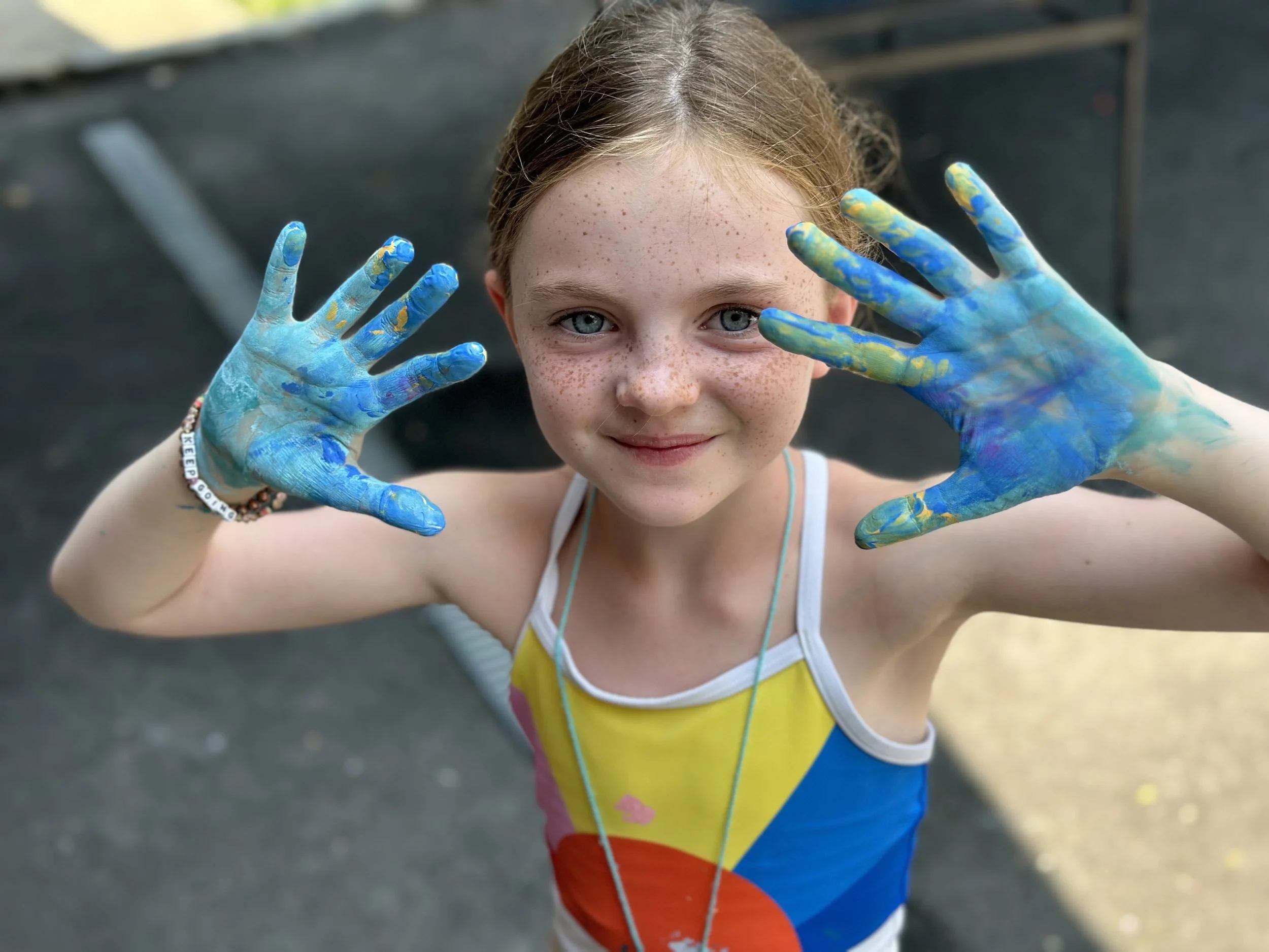 Young girl with light brown hair and freckles holding up her hands, covered in blue and yellow paint, smiling at the camera.