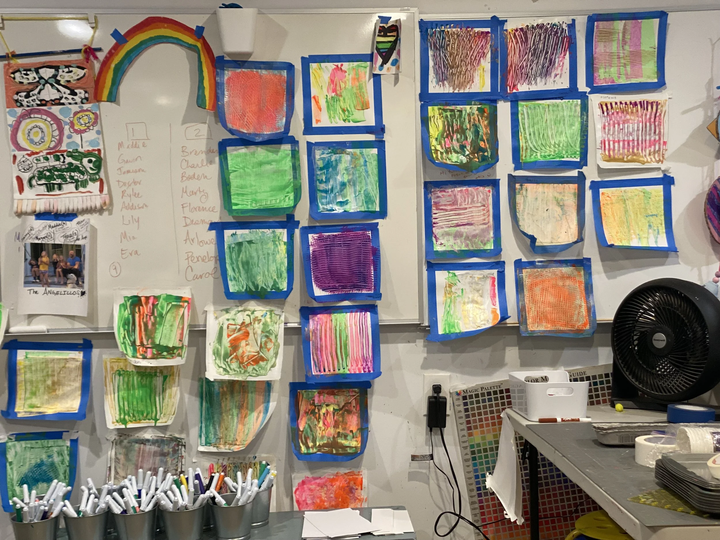Colorful children's artwork on a whiteboard, including paintings taped with blue painter's tape, a rainbow, collages, and a girl with a colorful shirt. Art supplies and a fan are also visible.