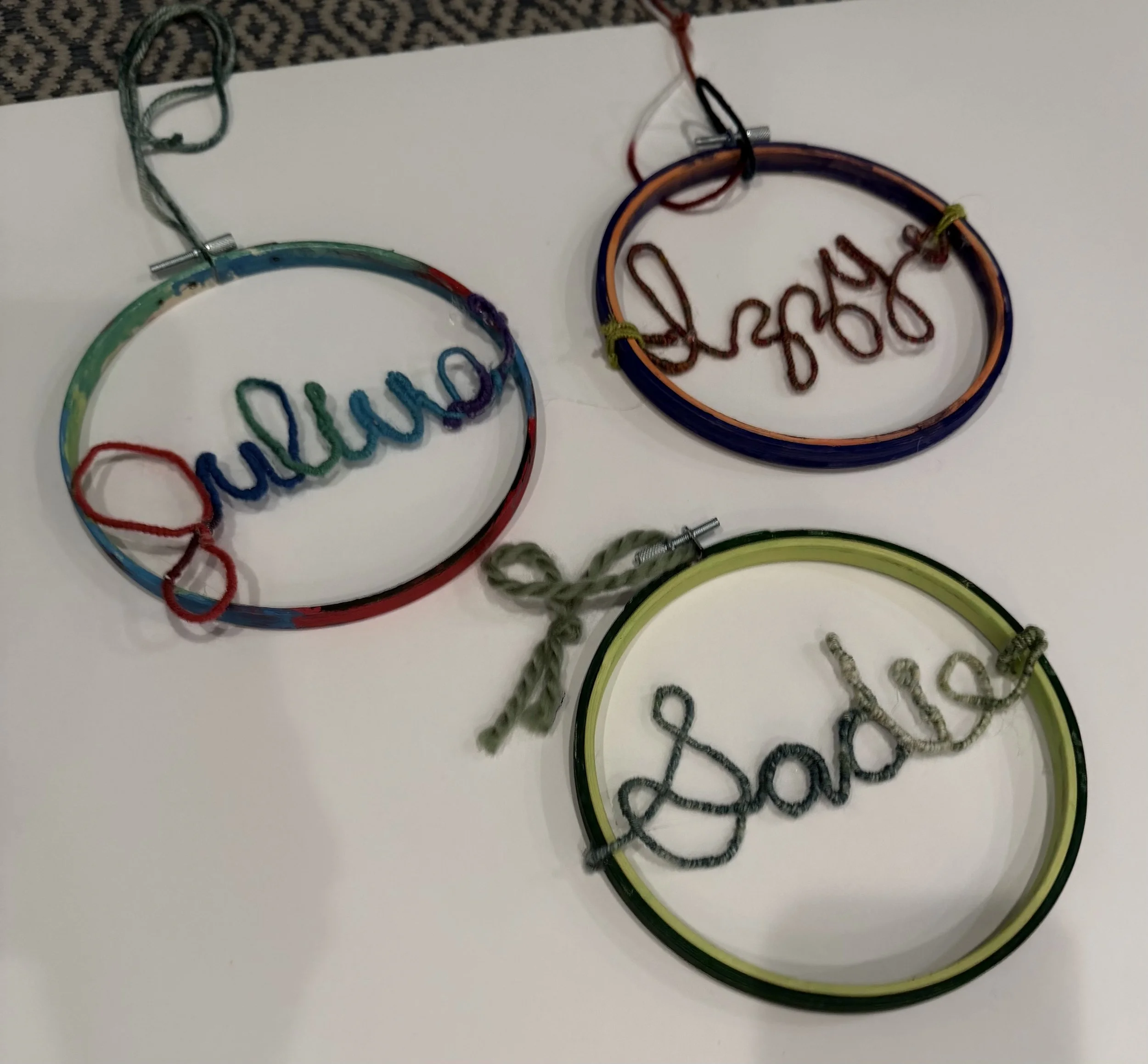 Three colorful circular wire name signs with the words "Juliana," "Lindsey," and "Lena" written in cursive, each tied with a small bow or knot, displayed on a white surface.