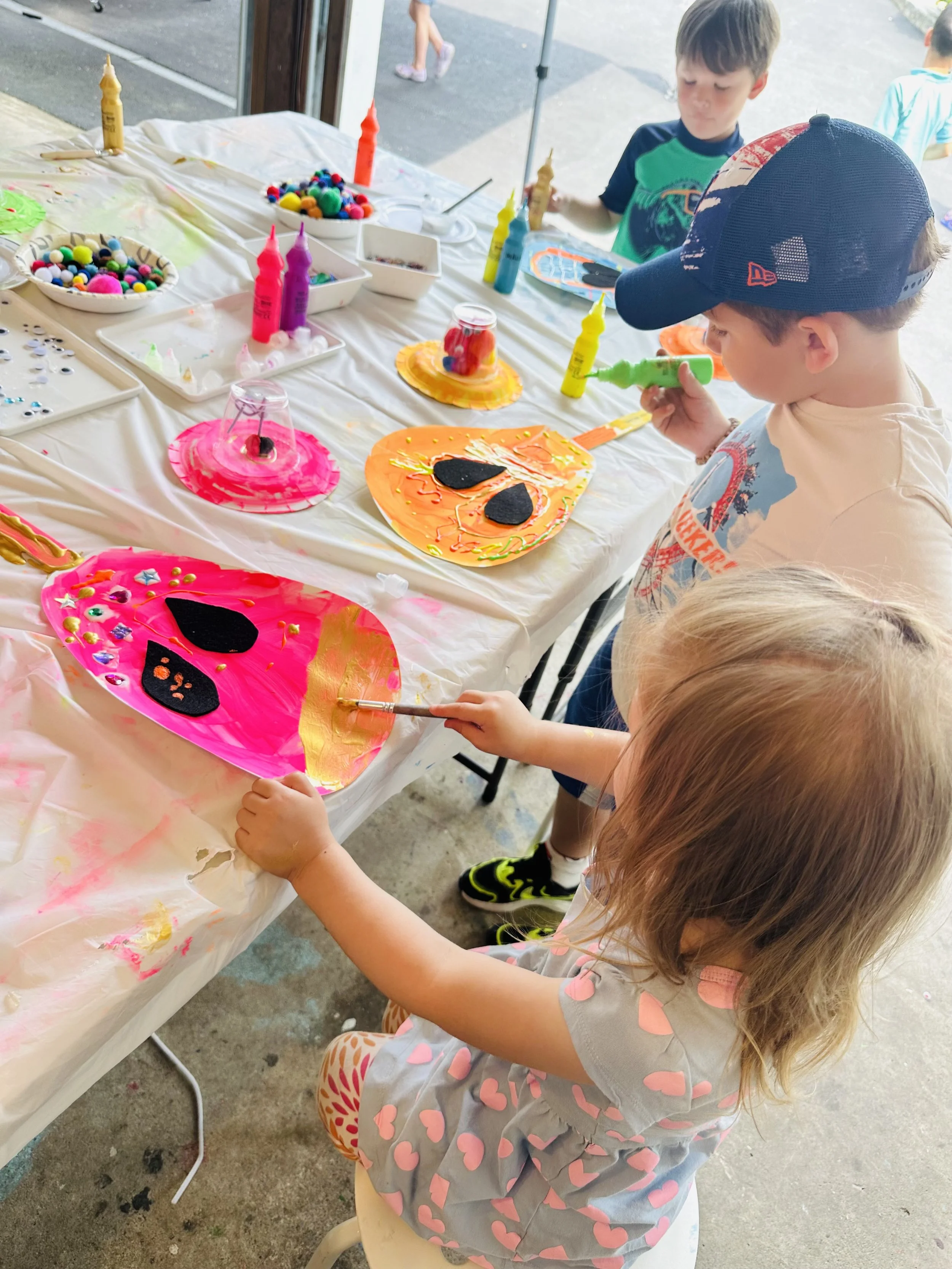 Children participating in a craft activity, decorating paper plates with paint, felt shapes, and small objects at a table covered with a plastic tablecloth.