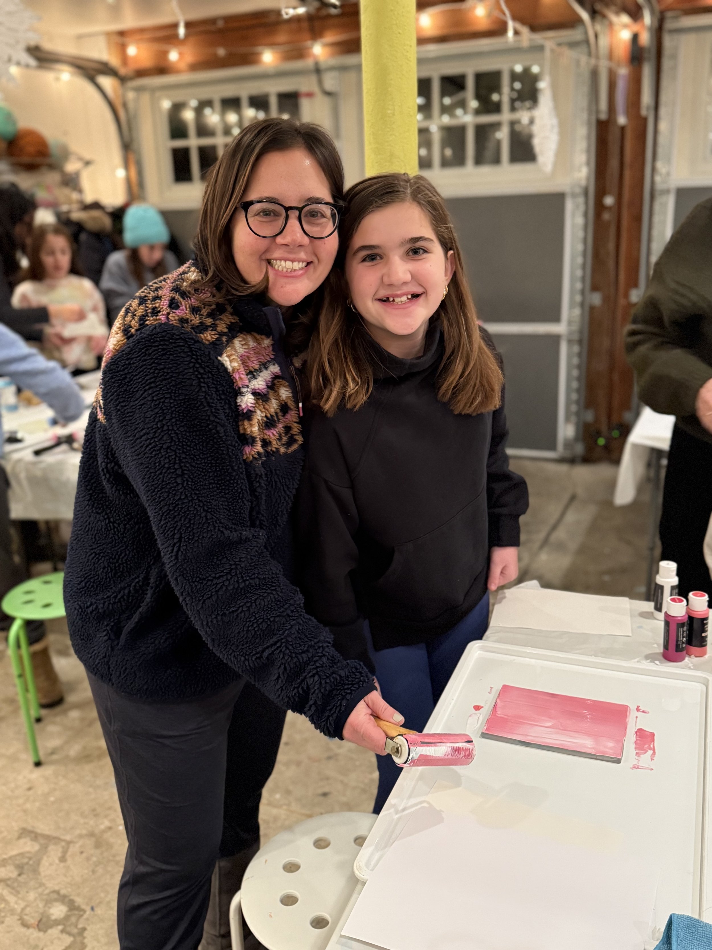 Two smiling females, a woman with glasses and a young girl, standing together in a festive indoor setting with Christmas decorations, painting on a small canvas.