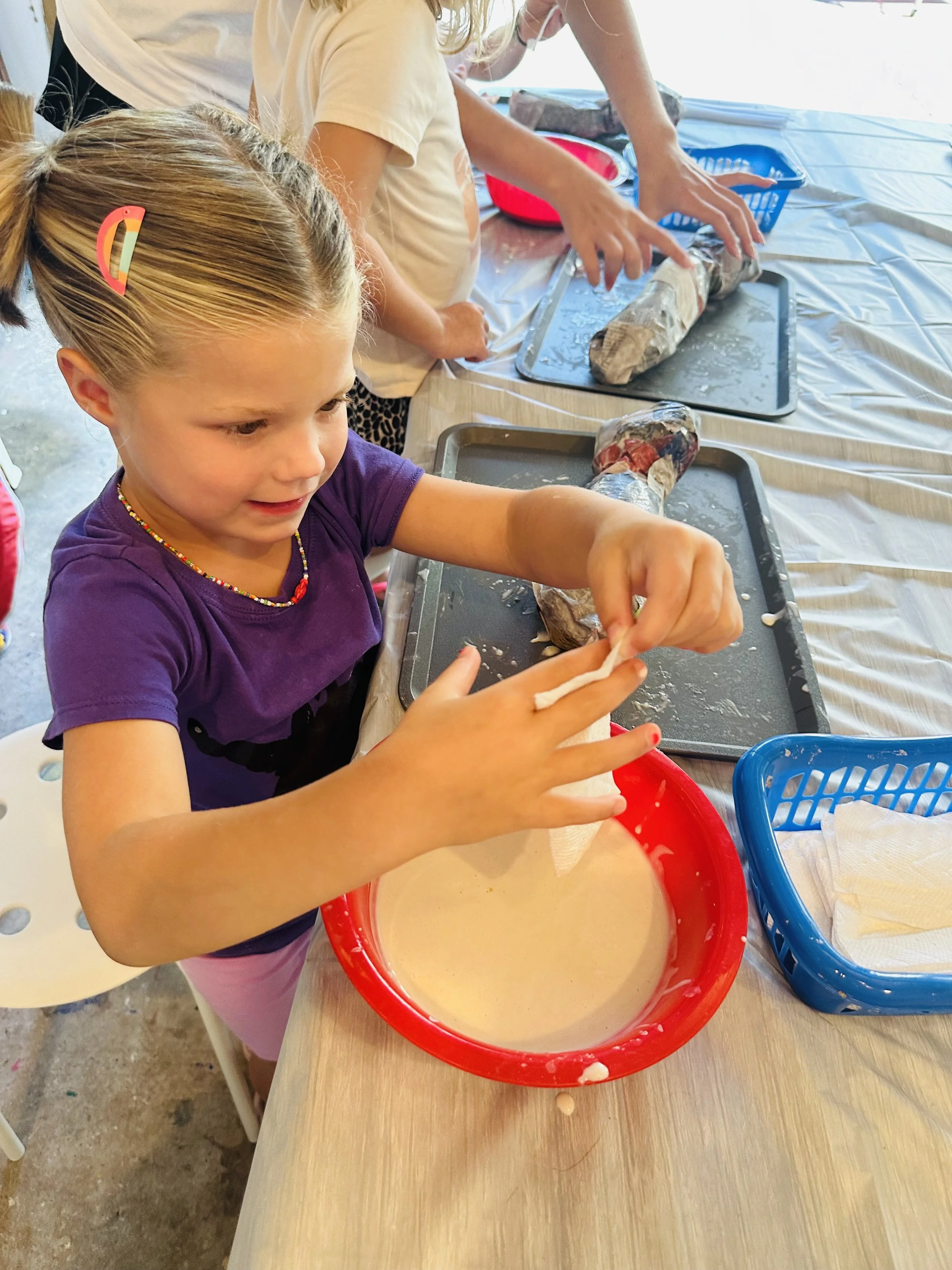 Young girl with hair in a ponytail wearing a purple shirt and beaded necklace, mixing a batter or dough in a red bowl, with other children and adults preparing food at a table.