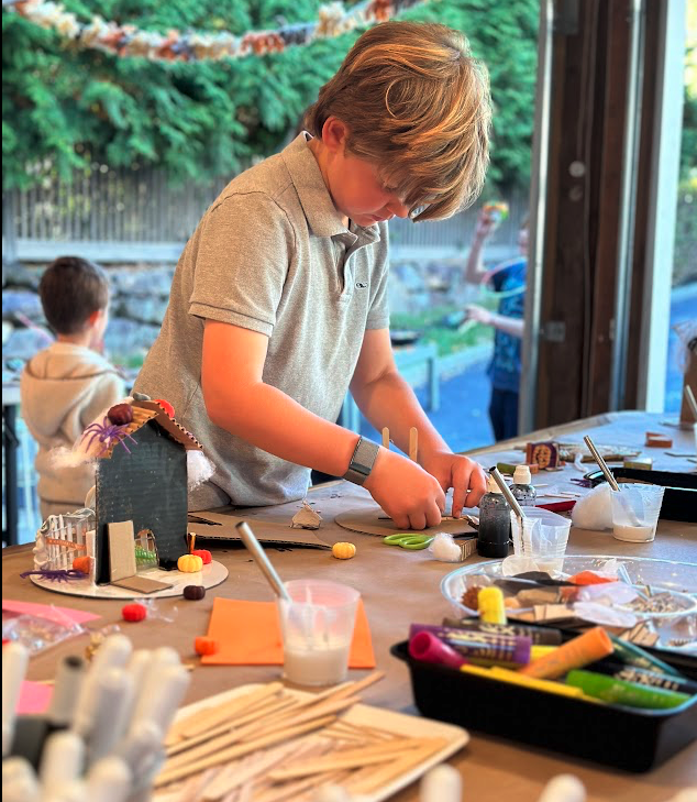 A young boy with blonde hair working on art projects at a table, surrounded by craft supplies, with a background of trees outside a window.