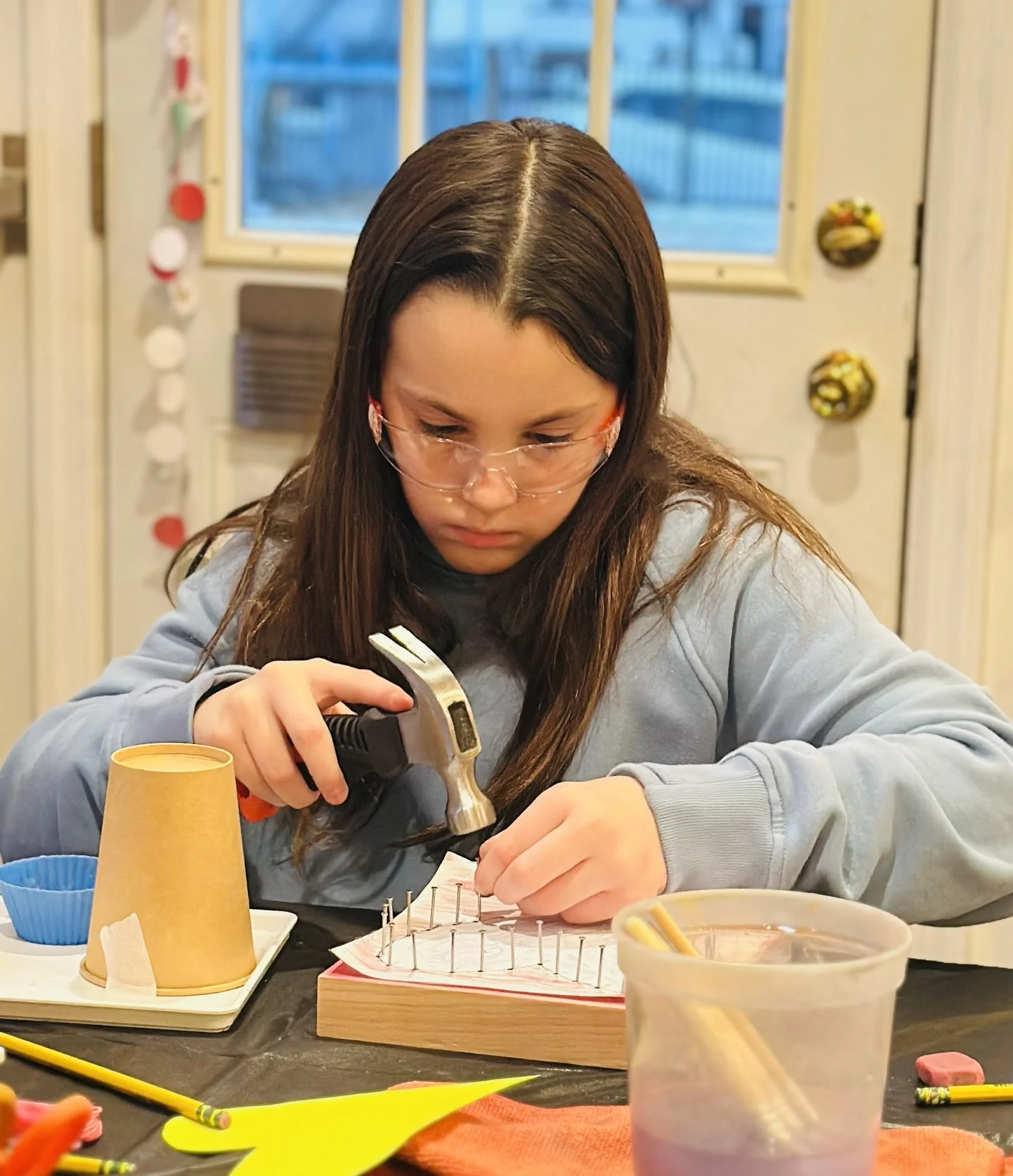A young girl wearing glasses is working on a craft project at a table, hammering nails into a wooden board.