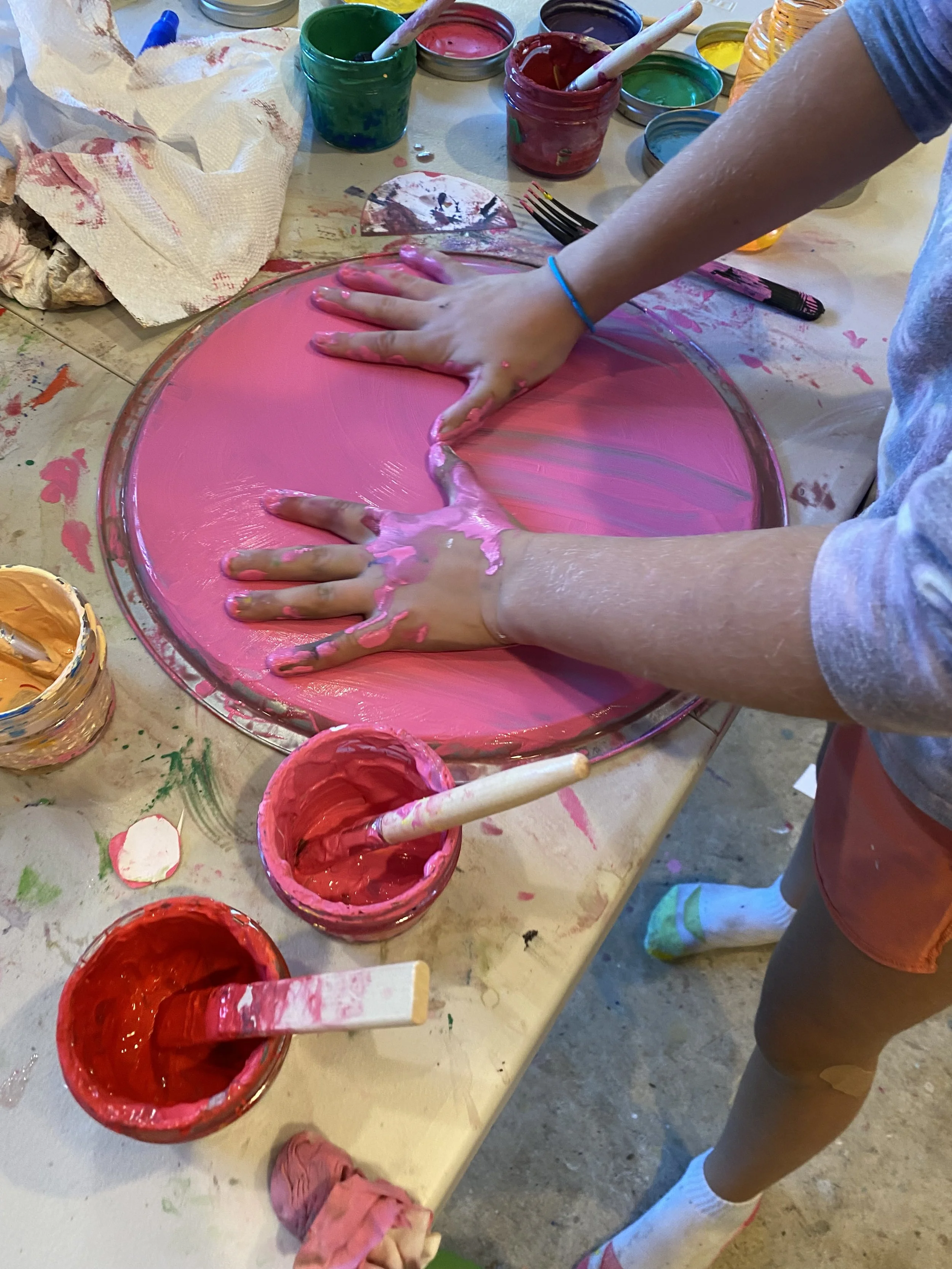 Child's hands spreading pink and purple paint on a circular surface at a messy arts and crafts table with various paint containers and brushes around.