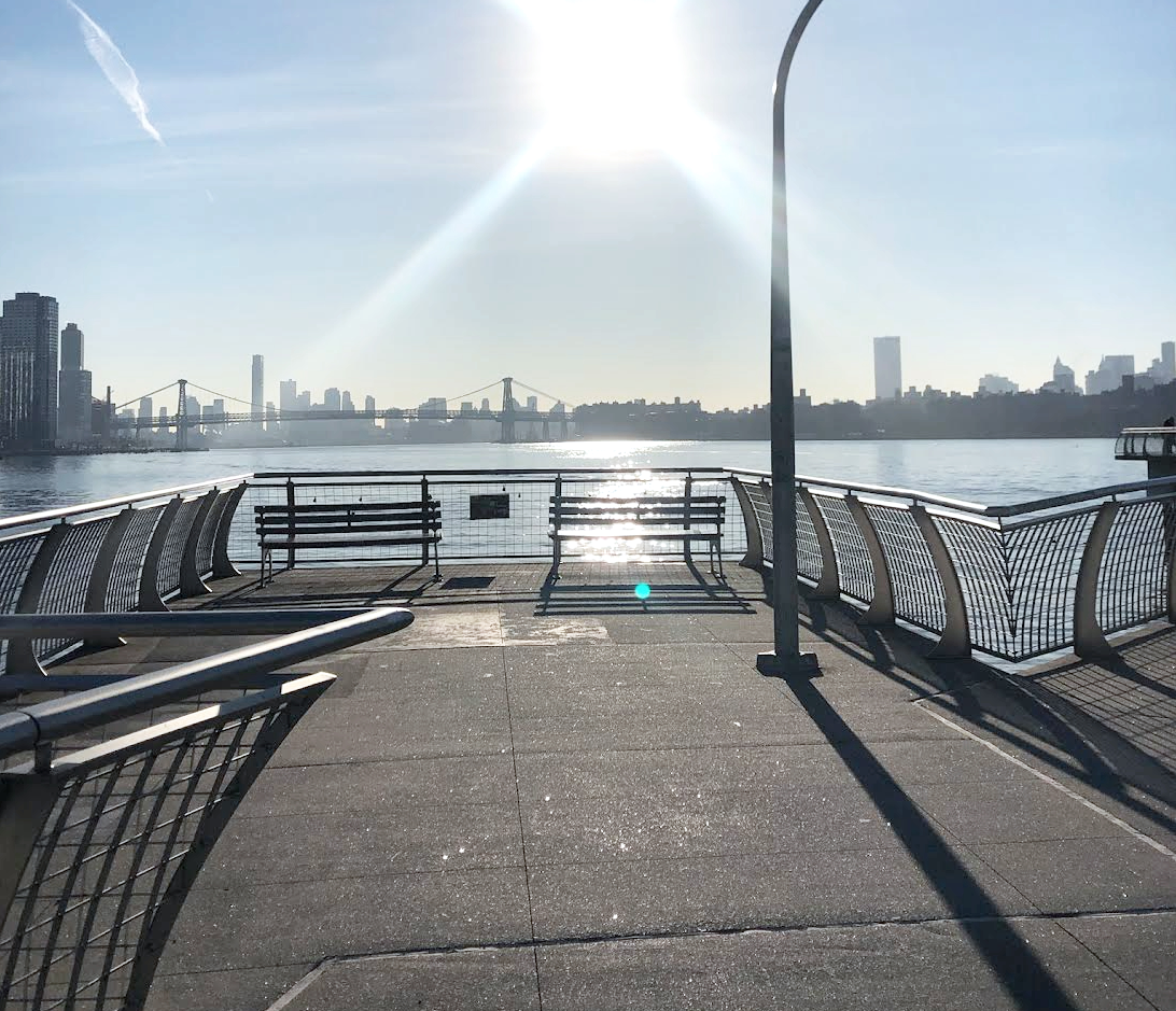 City skyline and bridge across a river seen from a waterfront promenade with benches and a streetlamp, sunlight creating reflections on the water.