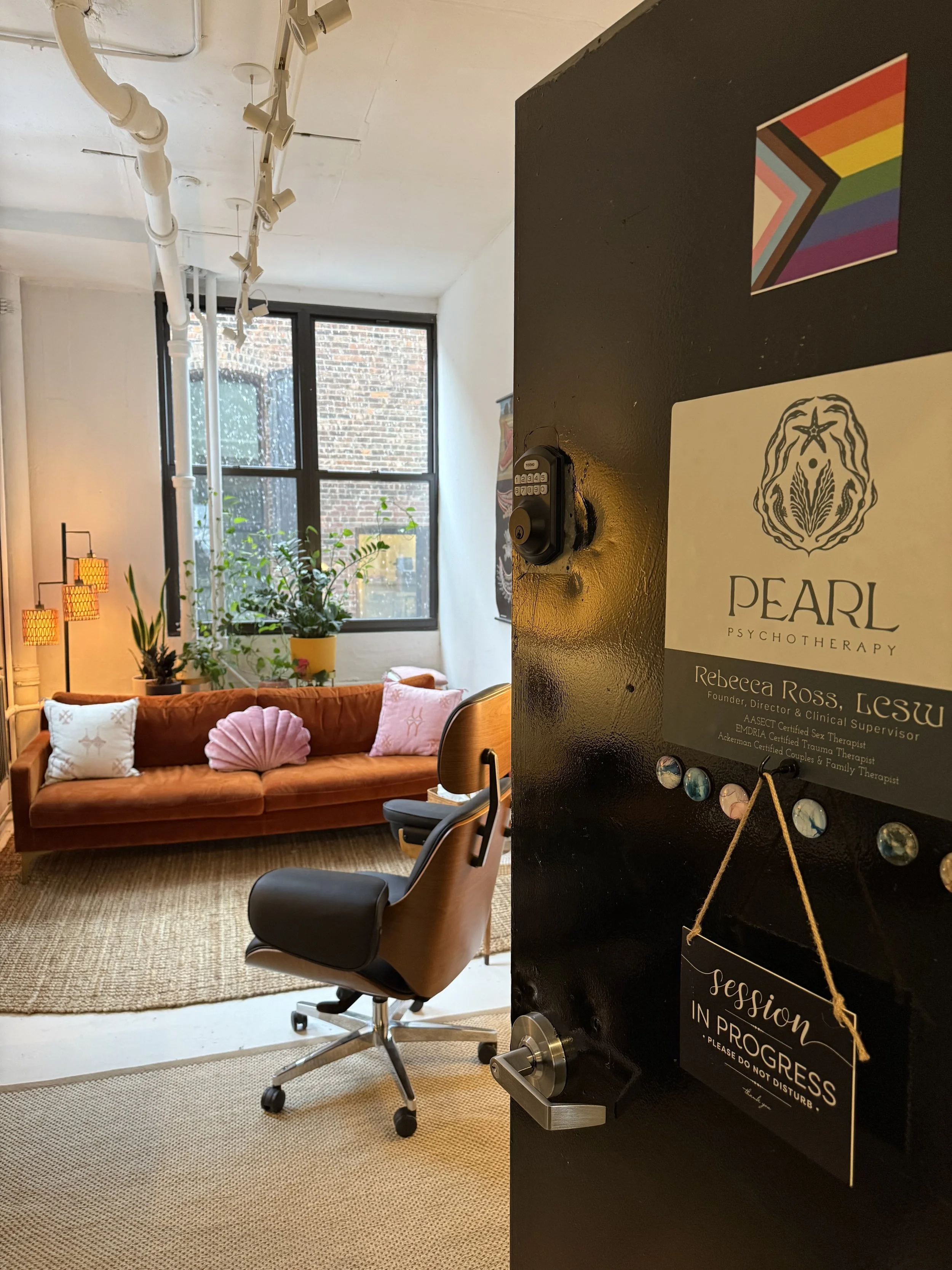 A cozy room with a burnt orange sofa decorated with white and pink pillows, a potted plant on a side table, a black office chair, and a large window with an exposed brick wall outside. There is a black partition with a colorful rainbow flag, a poster for Pearl Psychotherapy, and a session progress sign.