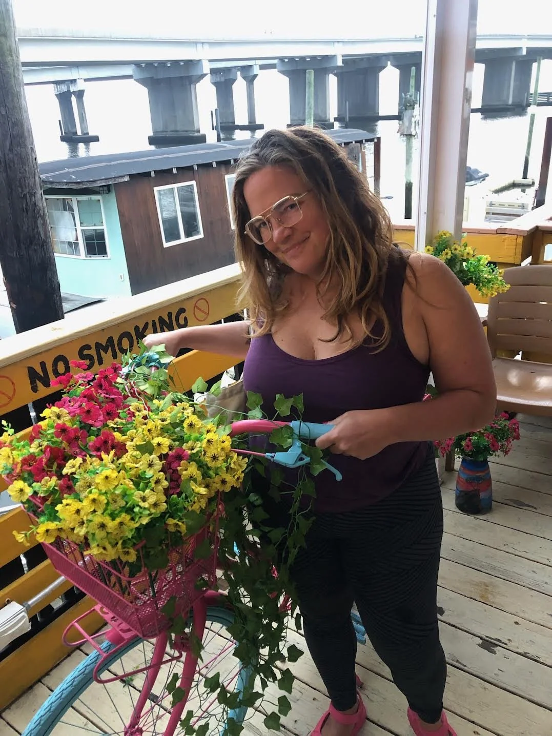 Woman with curly hair and glasses smiling while holding a pink bicycle with a basket of yellow and pink flowers, on a wooden deck near a body of water.
