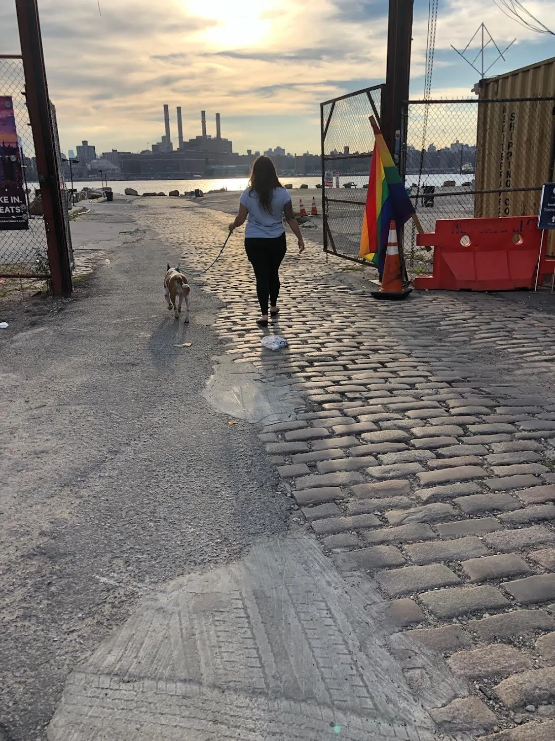 A woman walking a dog through a gated industrial area with cobblestone and asphalt ground, near a body of water with city buildings and power plant towers in the background during sunset.