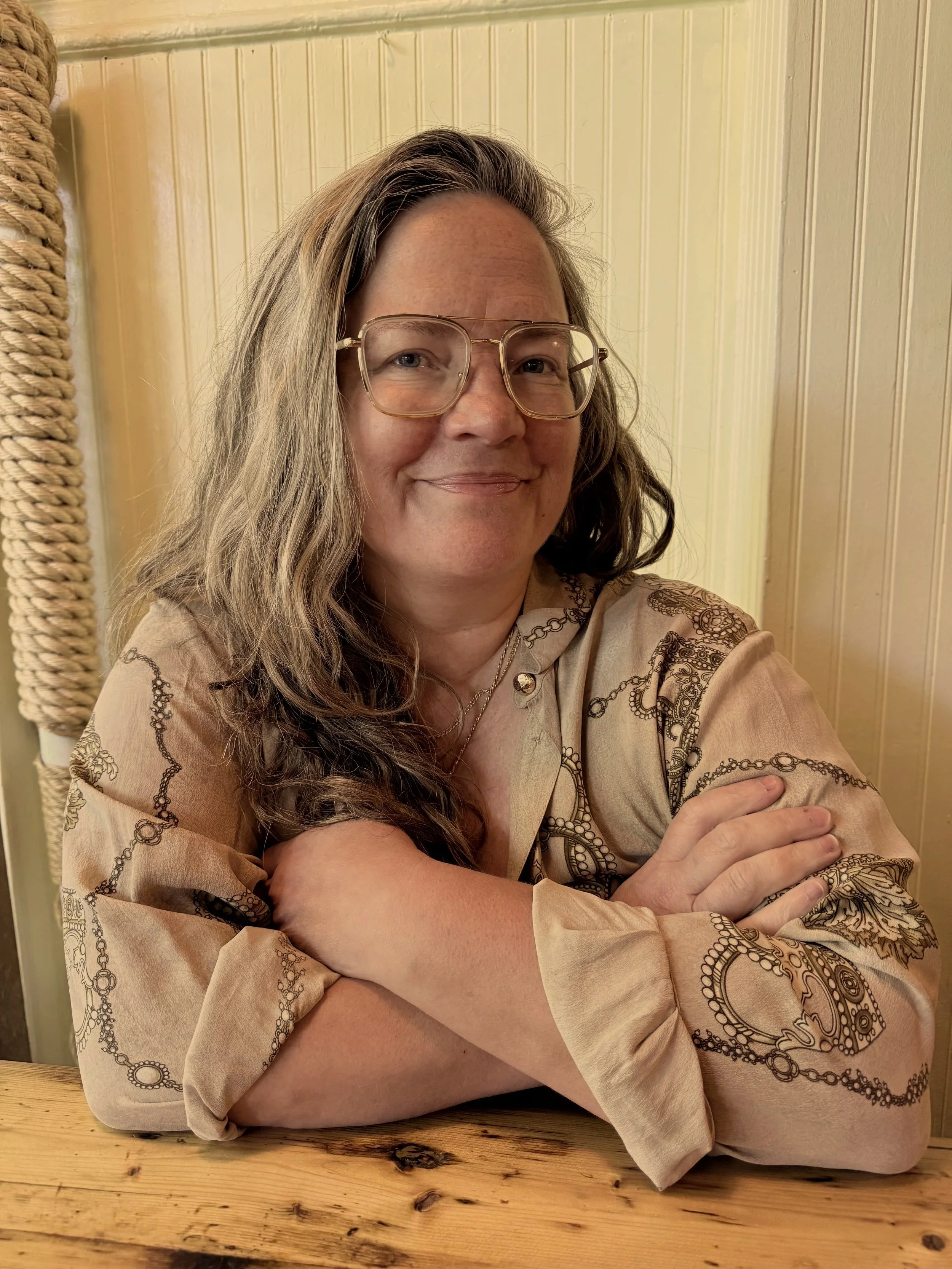 A woman with long, wavy hair and glasses, smiling, sitting at a wooden table in front of a beige wainscoted wall, wearing a patterned beige top.