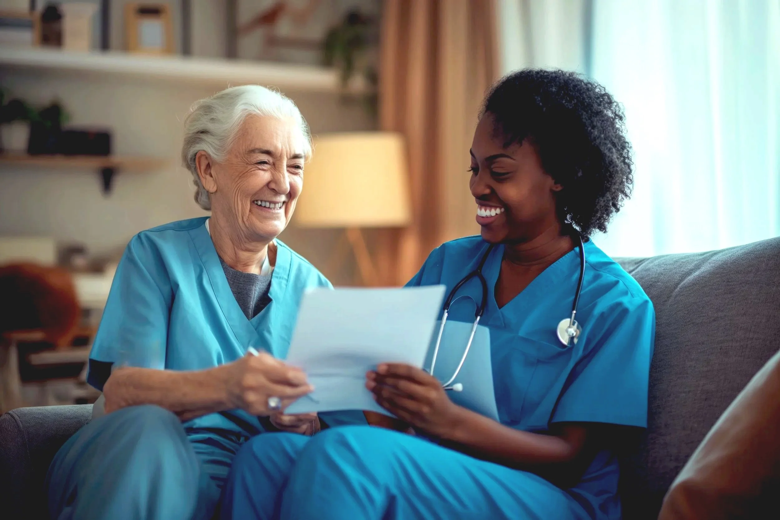 A senior woman and a female healthcare professional, both dressed in blue scrubs, sitting on a couch inside a cozy room. They are smiling and looking at a document held by the healthcare worker, sharing a moment of positive interaction.