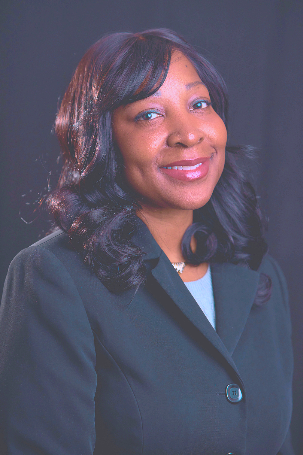 Professional woman with black hair, smiling, wearing a black blazer and light blue shirt, against a dark background.