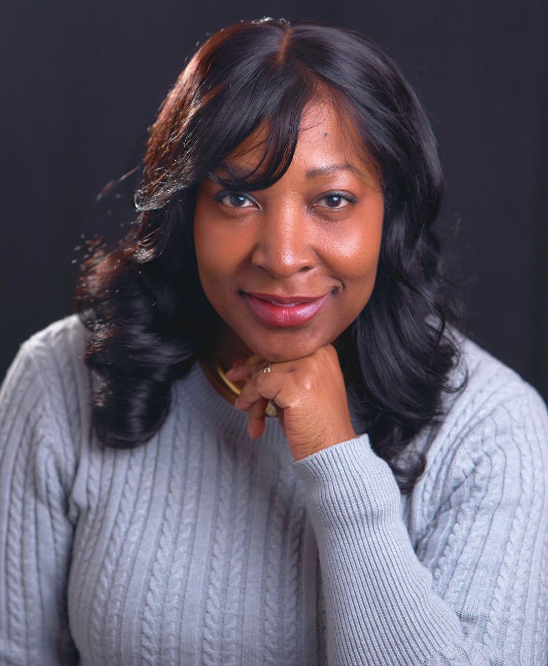 Portrait of a woman with black, wavy hair, wearing a gray sweater, smiling, with her chin resting on her hand, against a dark background.
