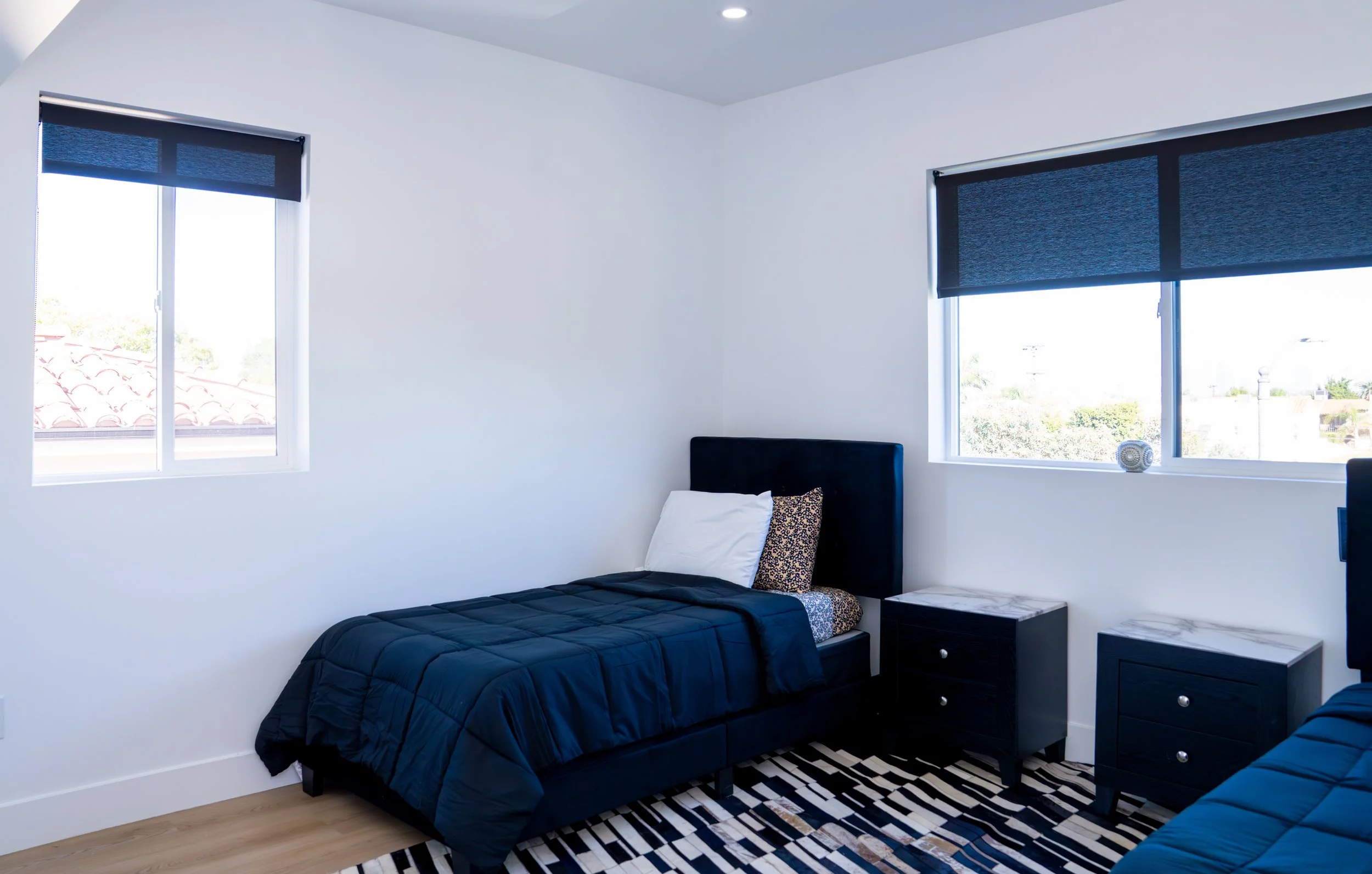 A bedroom with white walls, two windows with dark blue blinds, and two black twin beds with navy blue bedding. The bed on the left has a white pillow and a leopard print pillow. There are nightstands with books beside the beds. A black and white patt