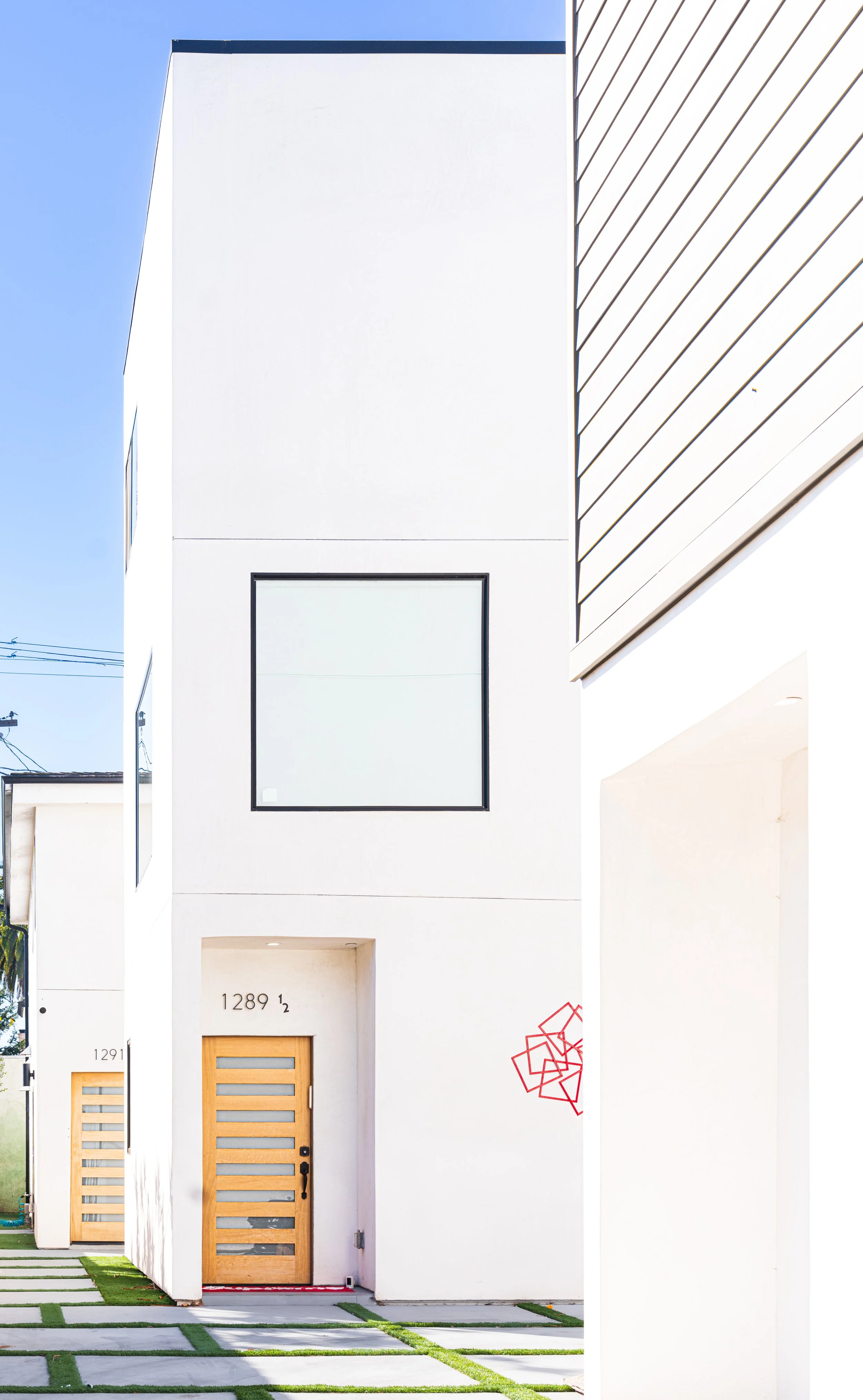 Modern white house with wooden front door, large window, and geometric landscaping in the foreground. Clear sky and neighboring buildings visible.